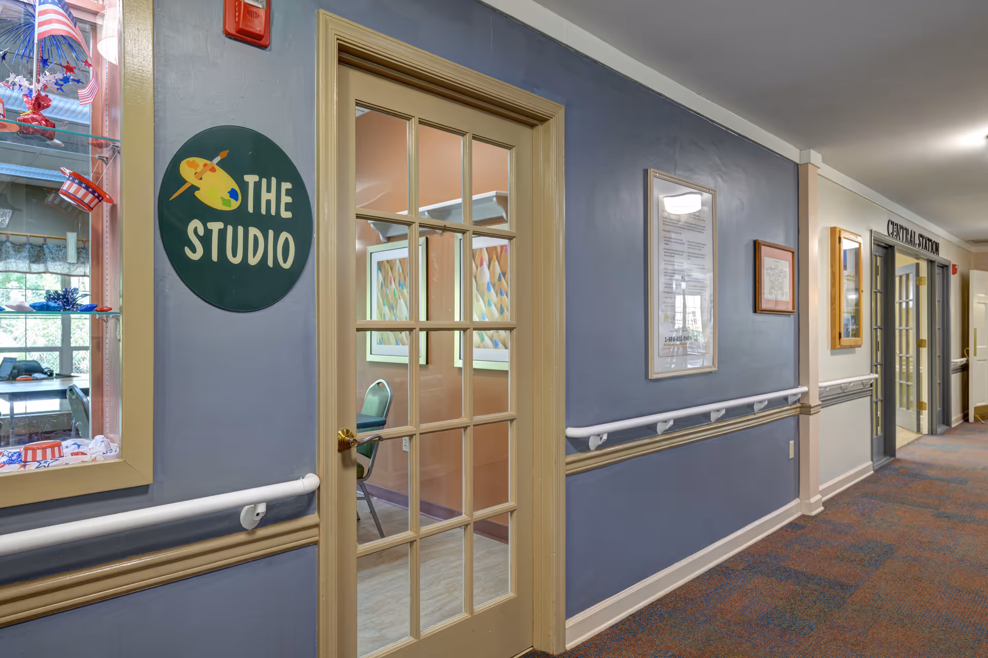 Interior hallway of a memory-care facility featuring a glass-paneled door labeled 'The Studio', handrails, and framed notices along the corridor.