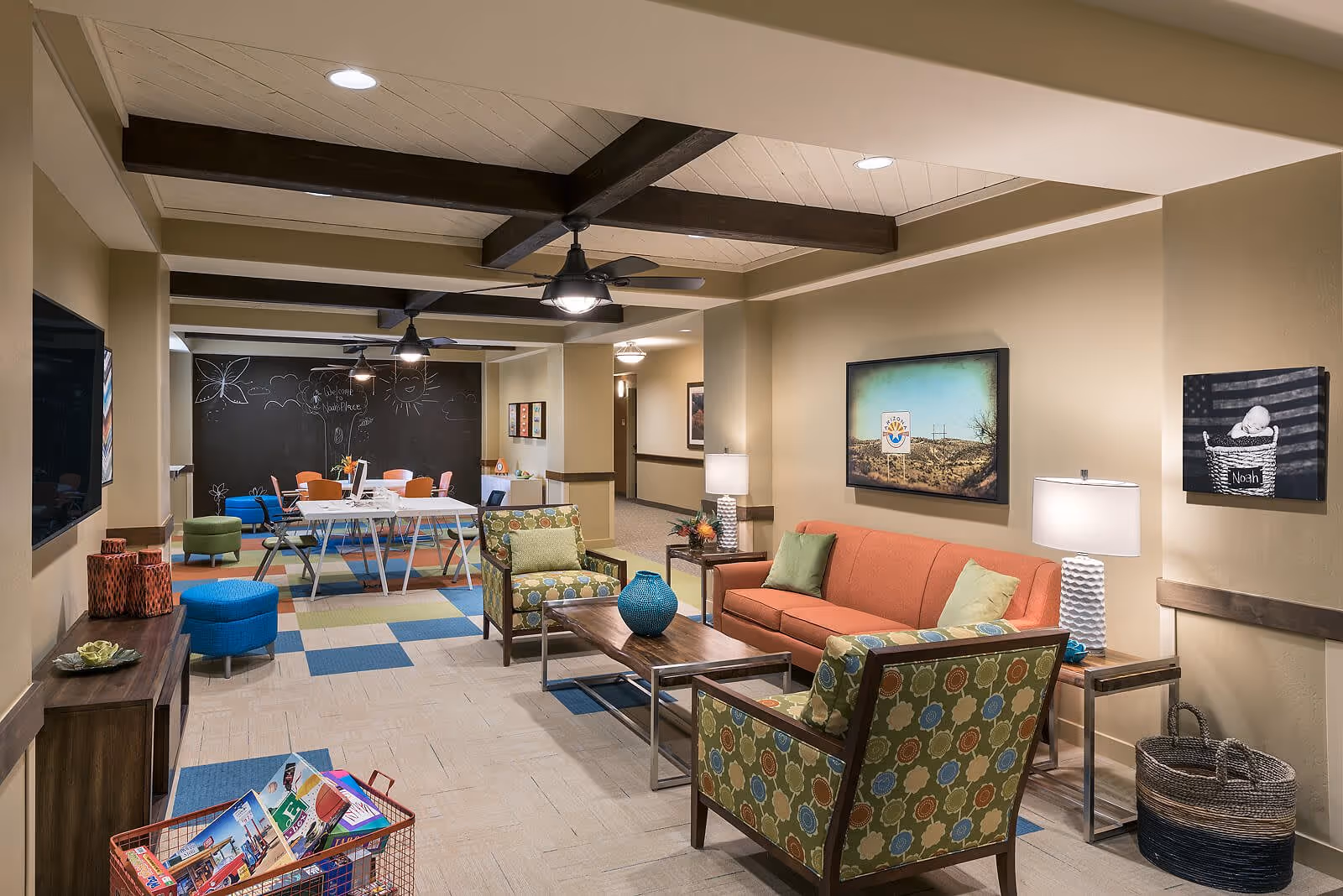 A cozy living room area in a senior living facility featuring an orange sofa, two patterned armchairs, a wooden coffee table with a blue vase, side tables with lamps, and colorful carpet tiles. In the background, there is a table with orange chairs and a large blackboard wall with drawings and the words 'Welcome Noah's Place'. The room has beige walls, ceiling fans with lights, and decorative wall art.