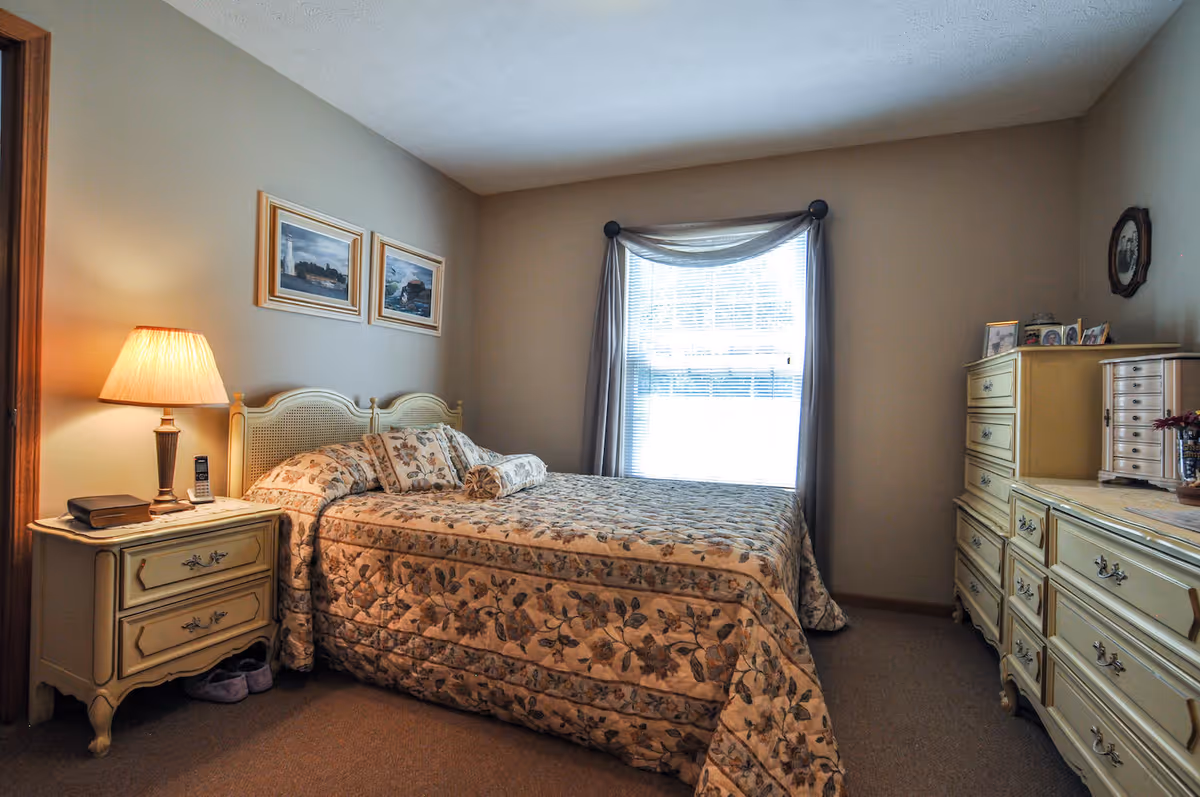 A cozy bedroom with a floral patterned bedspread on a double bed, a nightstand with a lamp and a phone, two framed pictures on the wall, a window with curtains, and a dresser with various items on top.