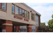 Exterior view of a building with a beige and brick facade, featuring large windows and the sign 'SUNSET HOME' prominently displayed above the entrance. Trees and a partly cloudy sky are visible in the background.