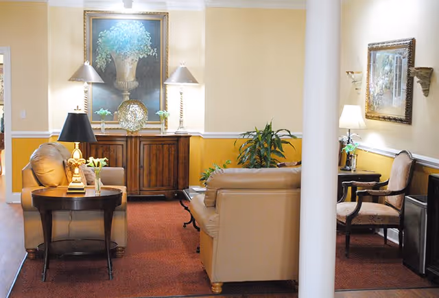 A cozy living room area with beige leather sofas, a wooden side table with a lamp and flowers, a wooden cabinet with two lamps and decorative items, a large framed painting of a vase with flowers on the wall, and a potted plant. The walls are painted yellow and cream, and there is a red carpet on the floor.