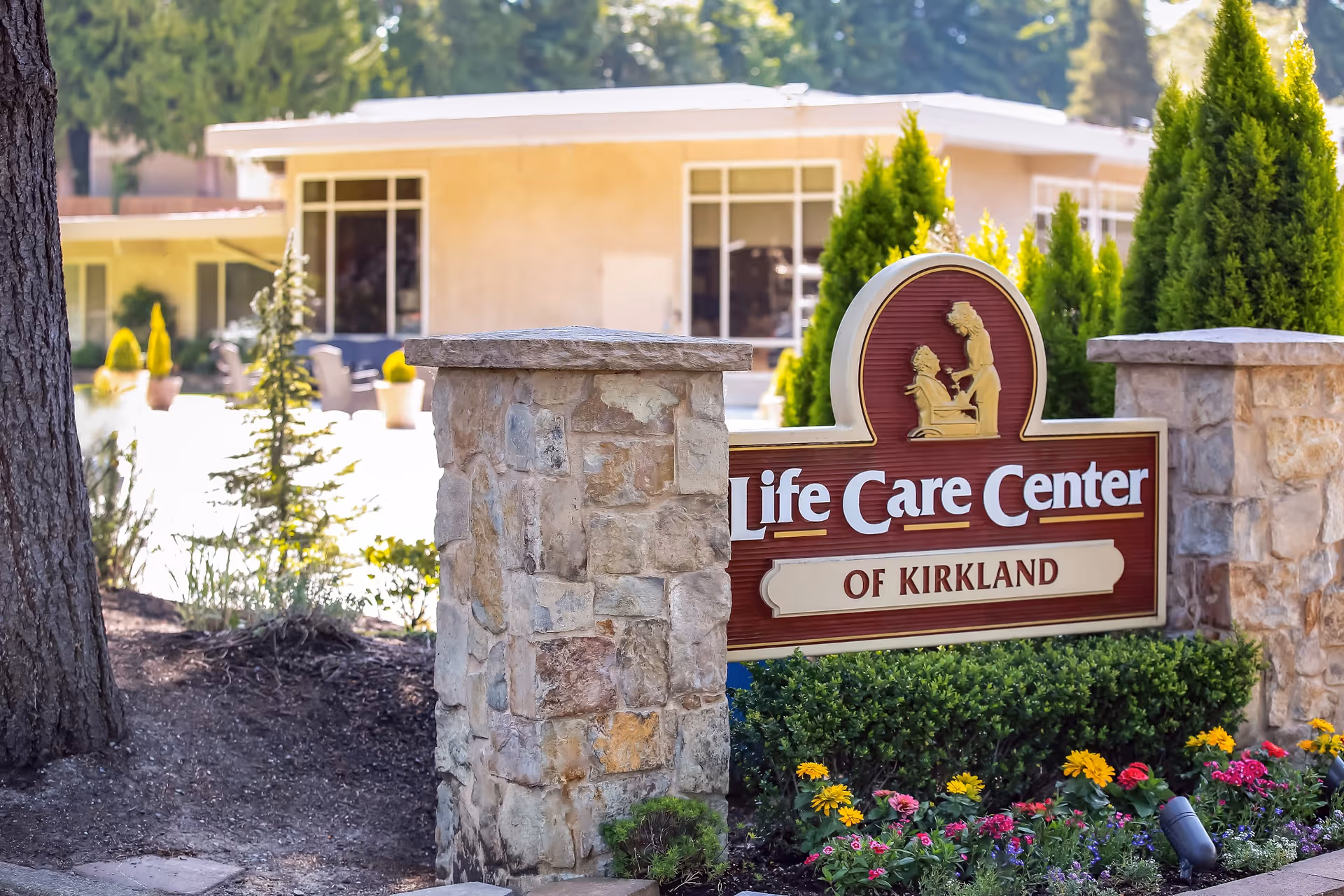 Stone pillars with a sign reading 'Life Care Center of Kirkland' surrounded by green shrubs and colorful flowers, with a building and outdoor seating area in the background.