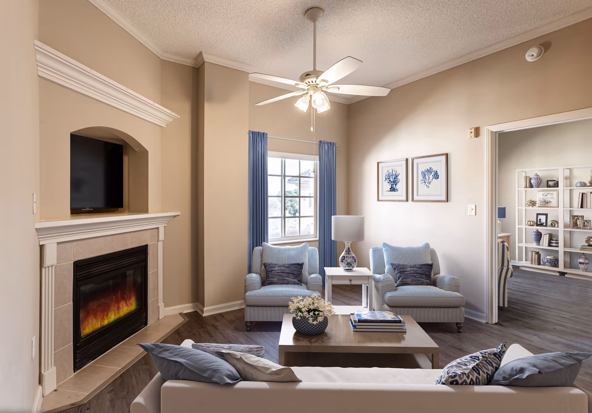 A cozy living room with a beige wall and ceiling fan. The room features a fireplace with a TV mounted above it, two striped armchairs with dark cushions, a white side table with a decorative lamp, a coffee table with books and a flower arrangement, and a white sofa with blue and patterned pillows. A window with blue curtains lets in natural light, and a doorway leads to another room with shelves displaying decorative items.