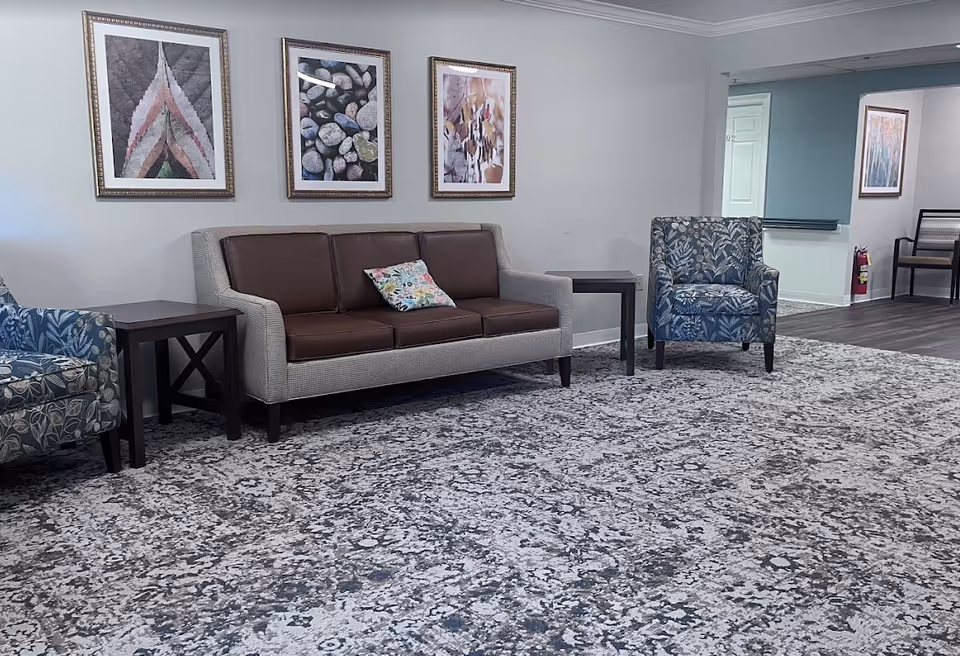 A seating area in a senior living facility with a gray and brown sofa, two patterned armchairs, two dark wooden side tables, and three framed abstract art pieces on the light gray wall above the sofa. The floor is covered with a large patterned carpet, and there is a hallway visible in the background with a fire extinguisher mounted on the wall.