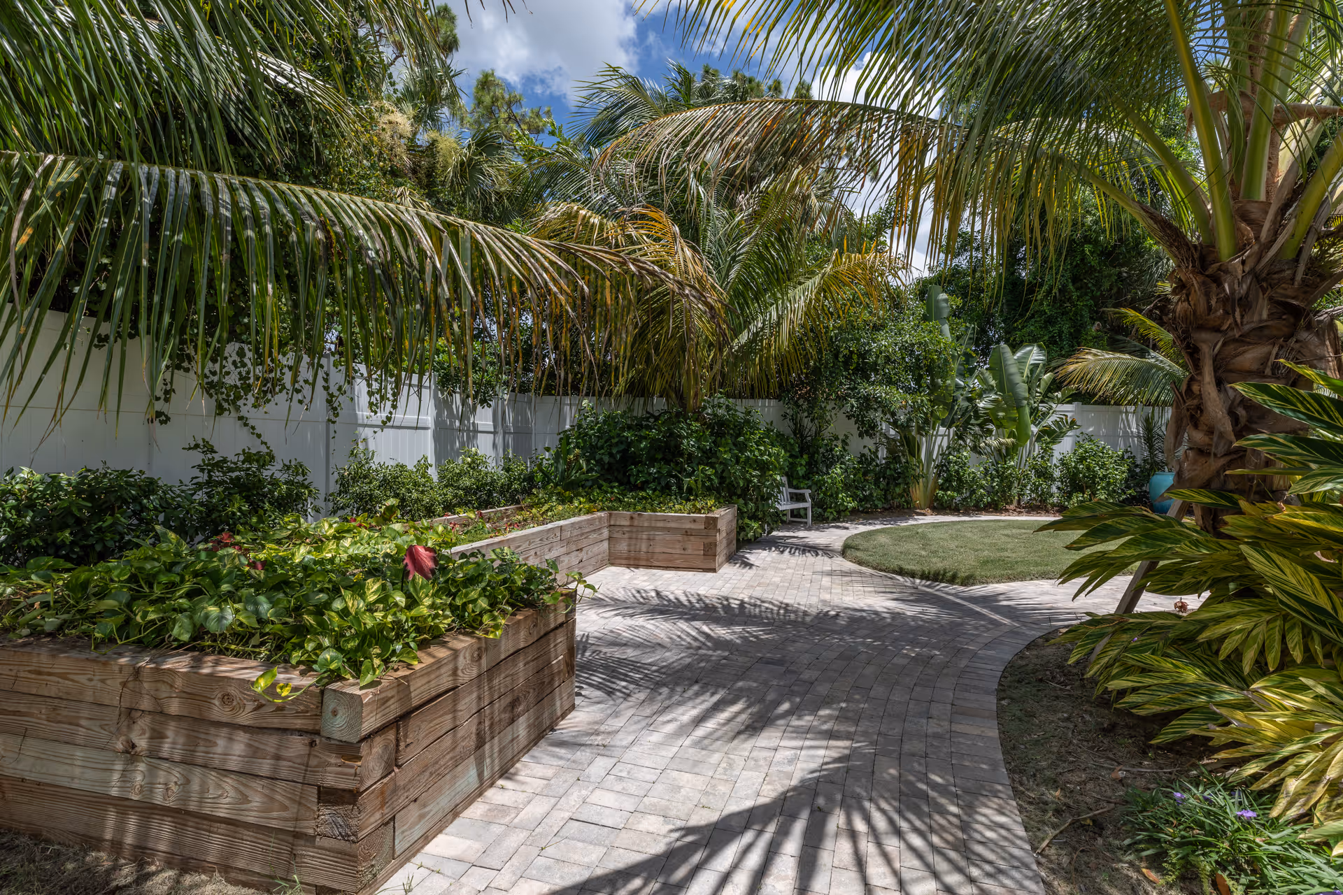 A paved garden path winds past raised wooden planter boxes and tropical palm trees in a sunny courtyard.