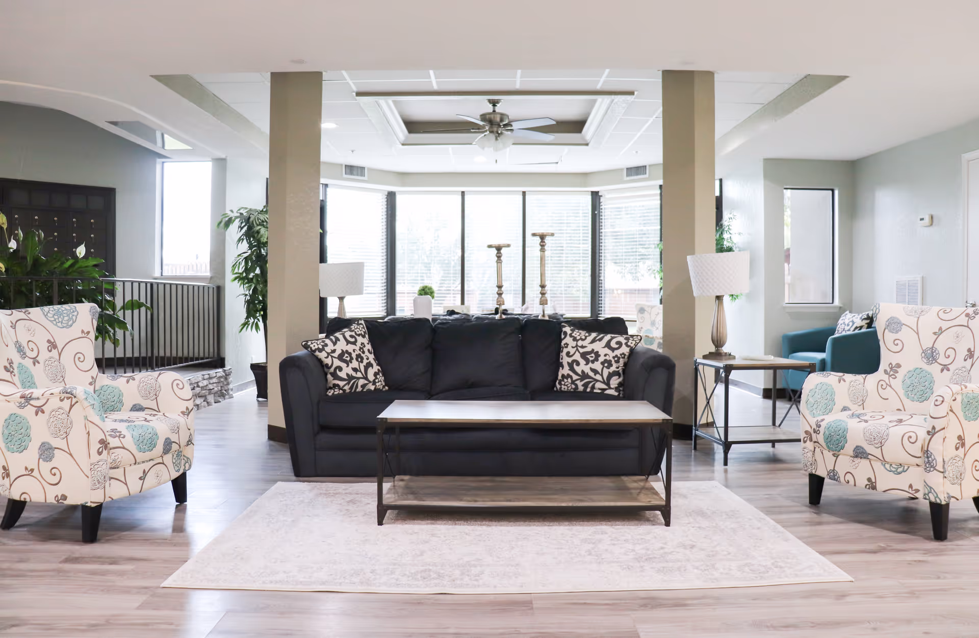 Bright communal living room with a dark sofa centered between patterned armchairs, a coffee table, lamps, and large windows.