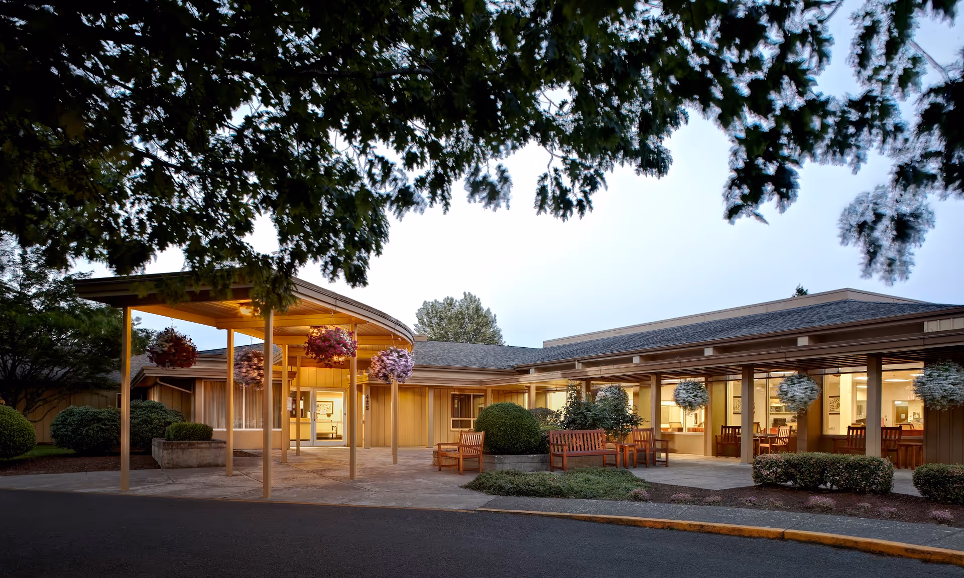 Exterior view of Avamere Riverpark of Eugene senior living facility at dusk, showing a covered entrance with hanging flower baskets, wooden benches, and well-maintained landscaping with bushes and trees.