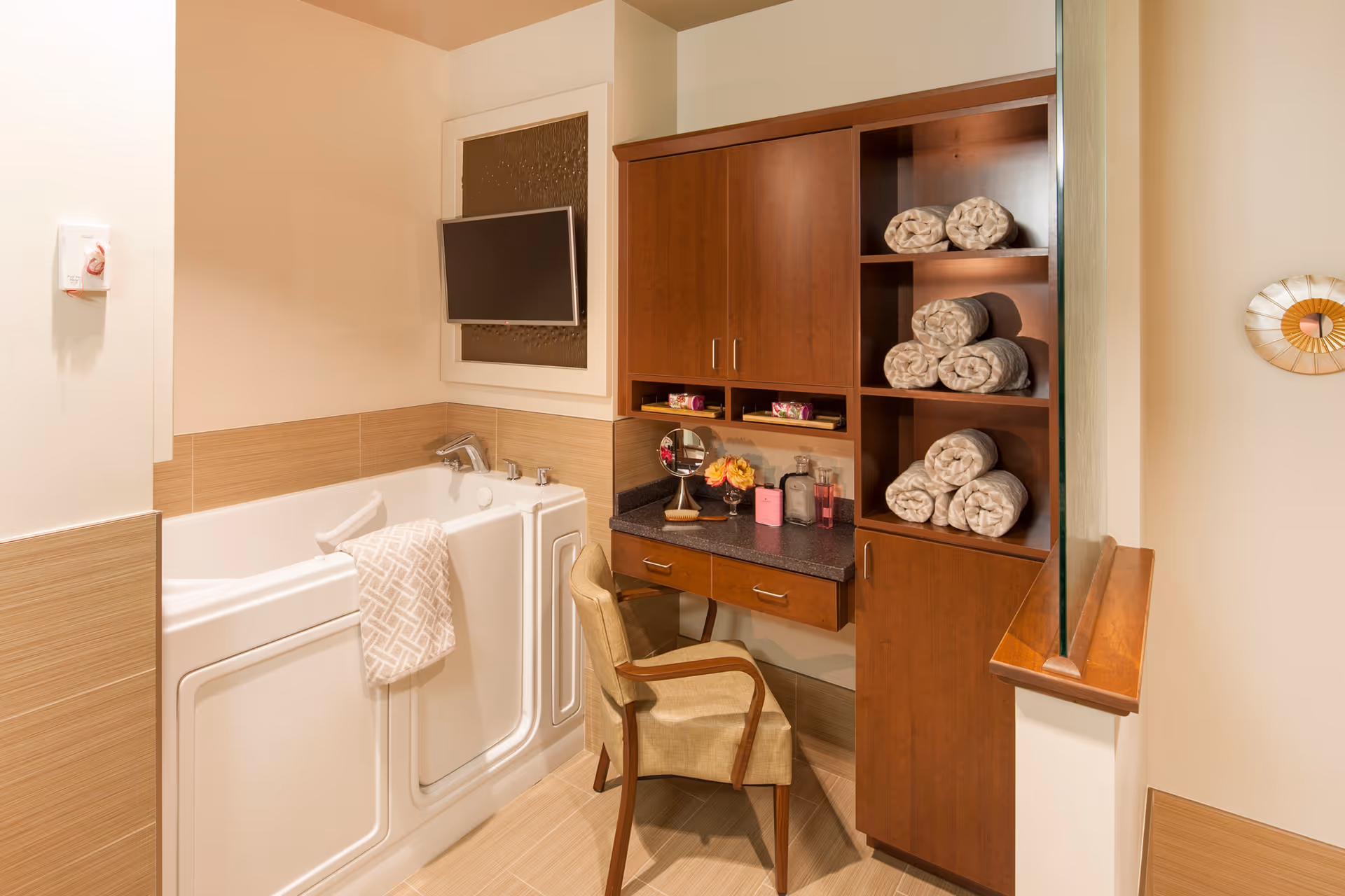 A bathroom featuring a walk-in bathtub with a towel draped over the side, a mounted flat-screen TV above the tub, and a wooden vanity with a chair. The vanity has a countertop with a small mirror, flowers, and toiletries. Next to the vanity is a wooden shelving unit with rolled towels neatly stacked.
