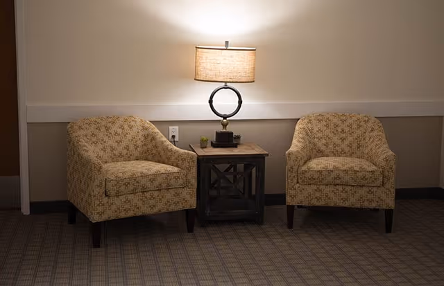 Two patterned armchairs flanking a wooden side table with a lit lamp in a carpeted interior seating area.