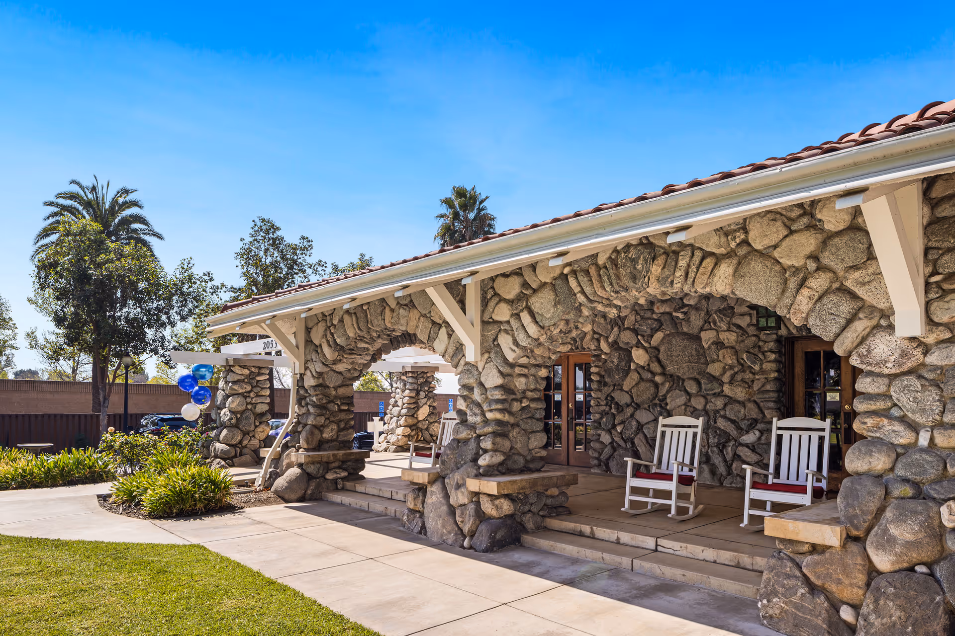 Stone building exterior with arched stone columns supporting a roof with red tiles. Two white rocking chairs with red cushions are placed on the porch. There are green plants and a manicured lawn in front, with a clear blue sky and some trees in the background.