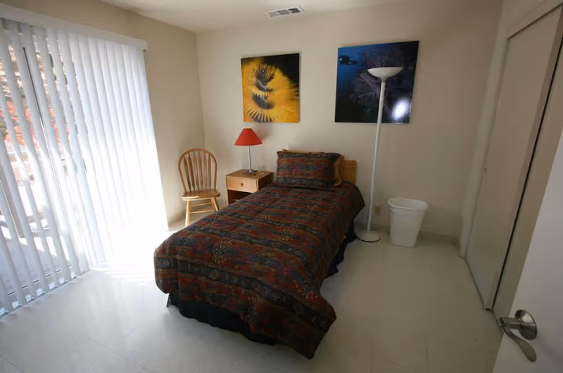 A small bedroom in a senior facility with a single bed, nightstand, chair, and posters.