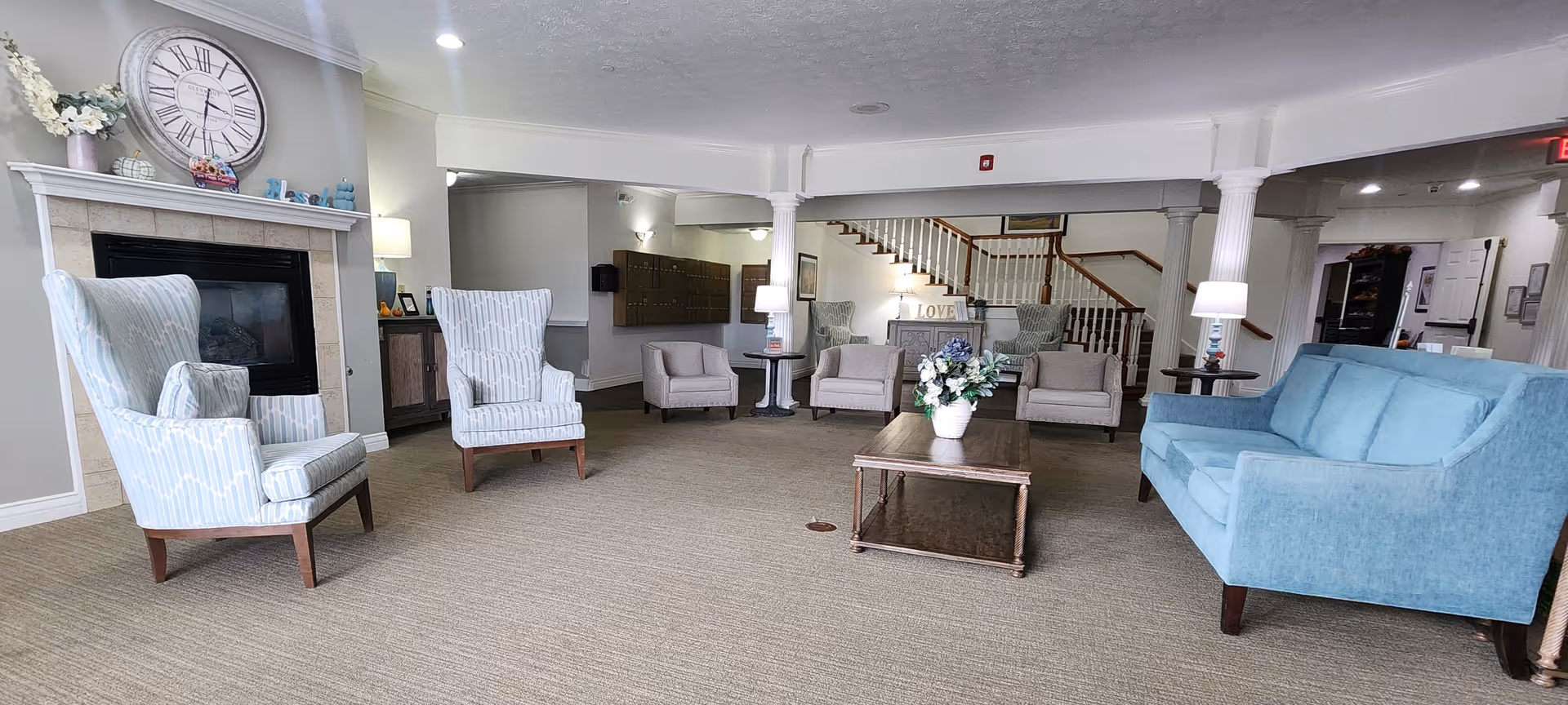 A spacious living room area in a senior living facility with light blue upholstered chairs and sofa arranged around a wooden coffee table with a flower vase. There is a fireplace with a large clock above it on the left side, and a staircase with wooden railings in the background. The room is well-lit with ceiling lights and table lamps, and has neutral-colored walls and carpeted flooring.