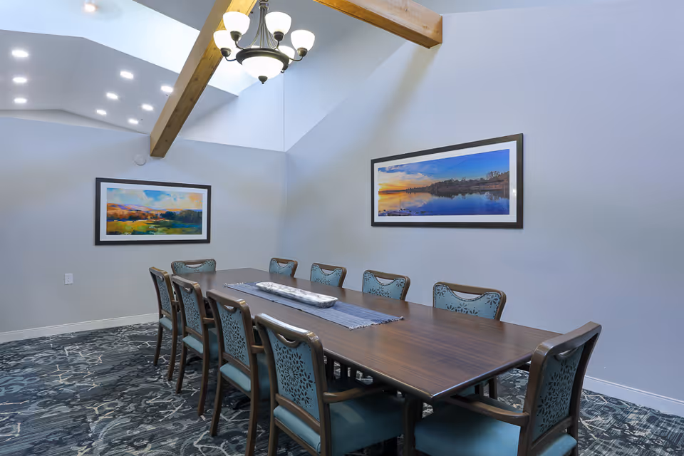 Long communal dining table with upholstered chairs beneath a skylight and chandelier, with framed landscape artwork on the walls.