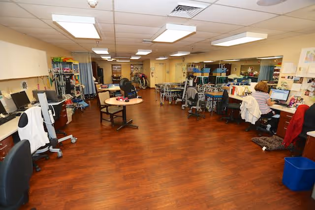 A spacious room with wooden flooring and fluorescent ceiling lights, featuring multiple desks and chairs arranged along the walls and in the center. A person is seated at a desk on the right side working on a computer. The room contains various office and medical equipment, suggesting a functional workspace within a healthcare facility.