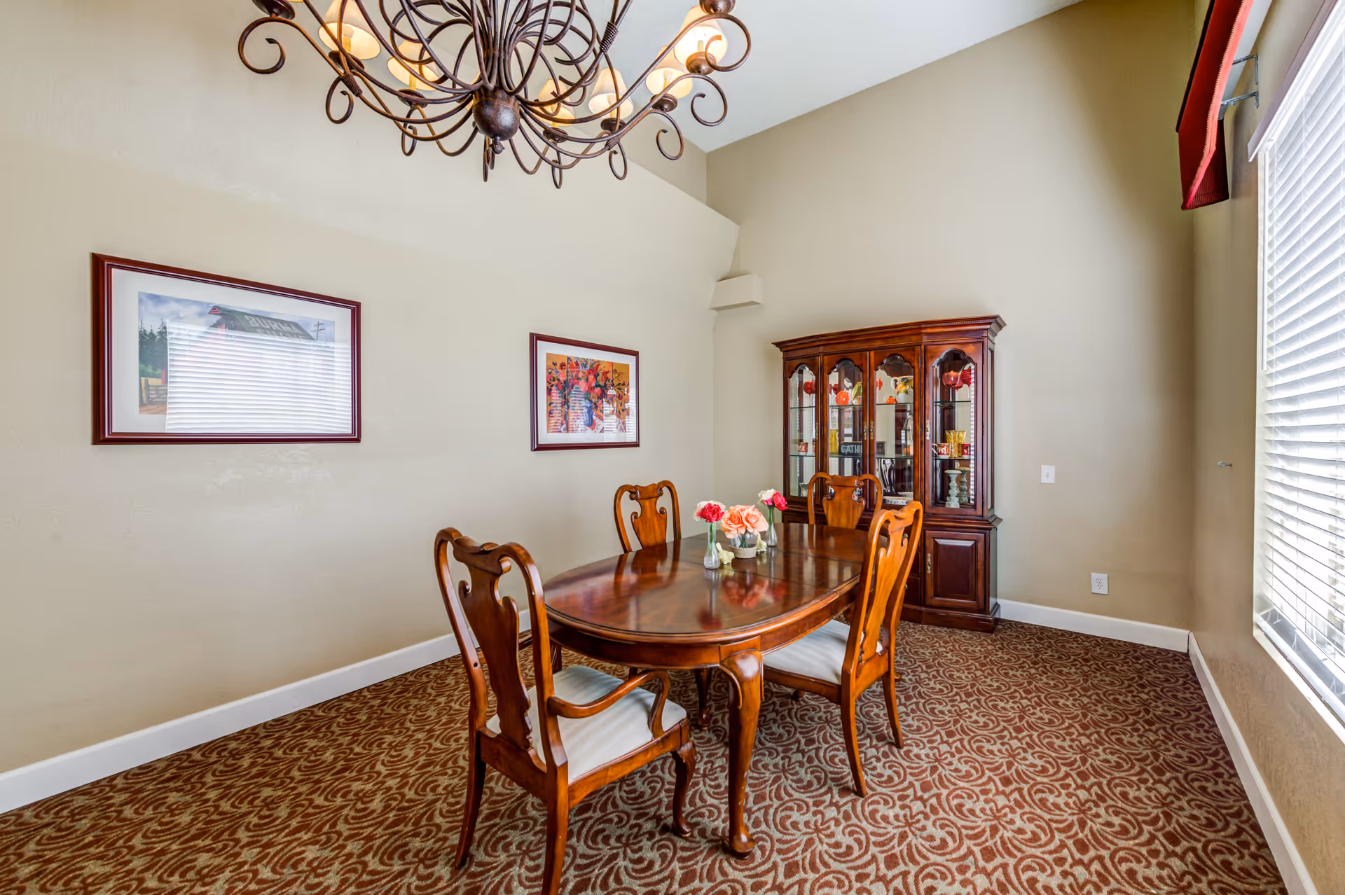 A dining room with a wooden dining table and four matching chairs with white cushions. The table has three small vases with pink and red flowers. There is a wooden china cabinet with glass doors displaying decorative items. Two framed pictures hang on the beige wall. A large window with white blinds and a red valance lets in natural light. A decorative chandelier hangs from the ceiling. The floor is covered with a patterned carpet.