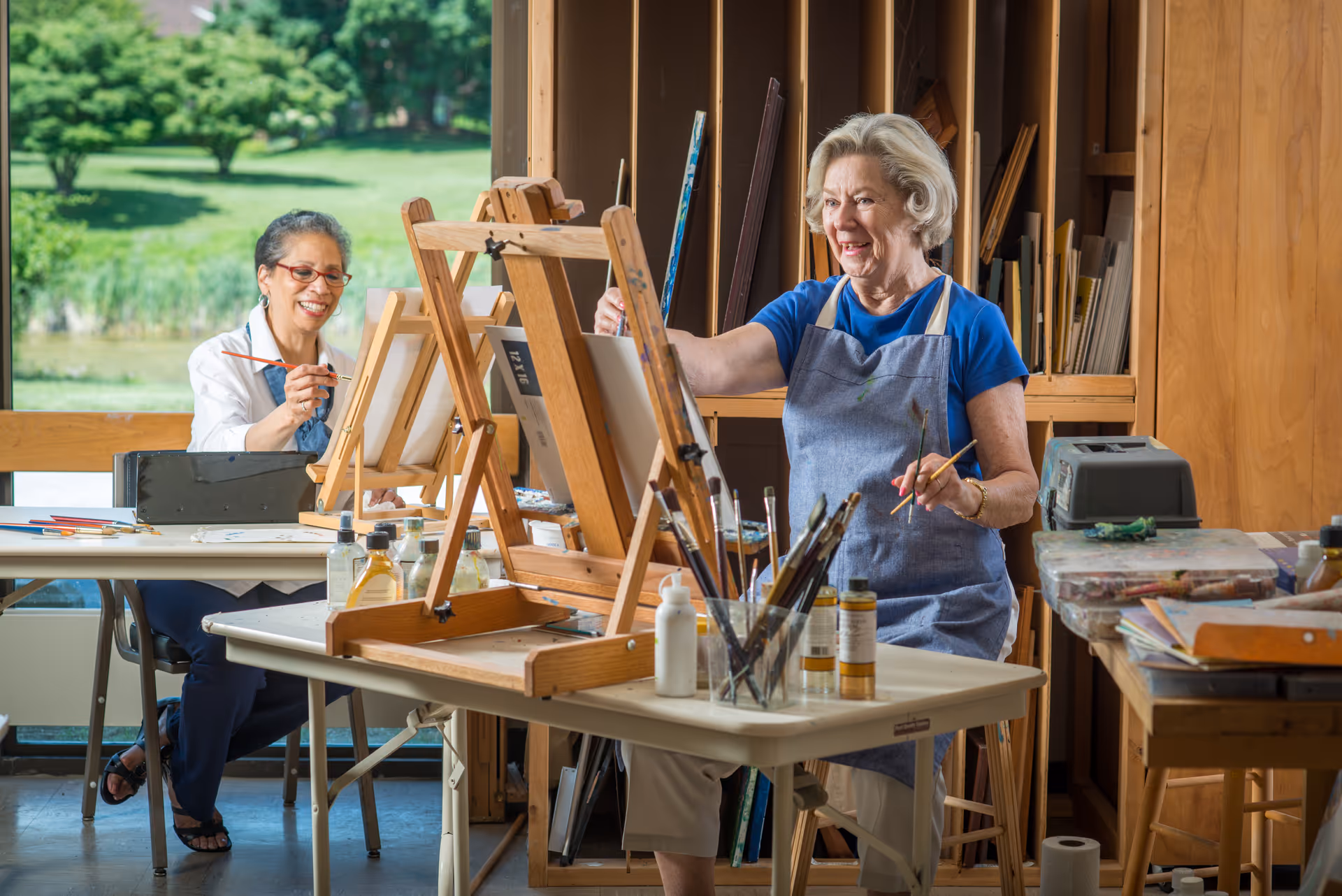 Two elderly women painting on easels in a bright art studio with large windows showing a green outdoor landscape. One woman is seated and smiling while painting, and the other is standing and also smiling, wearing a blue apron. The room contains art supplies, brushes, and wooden shelves with canvases.