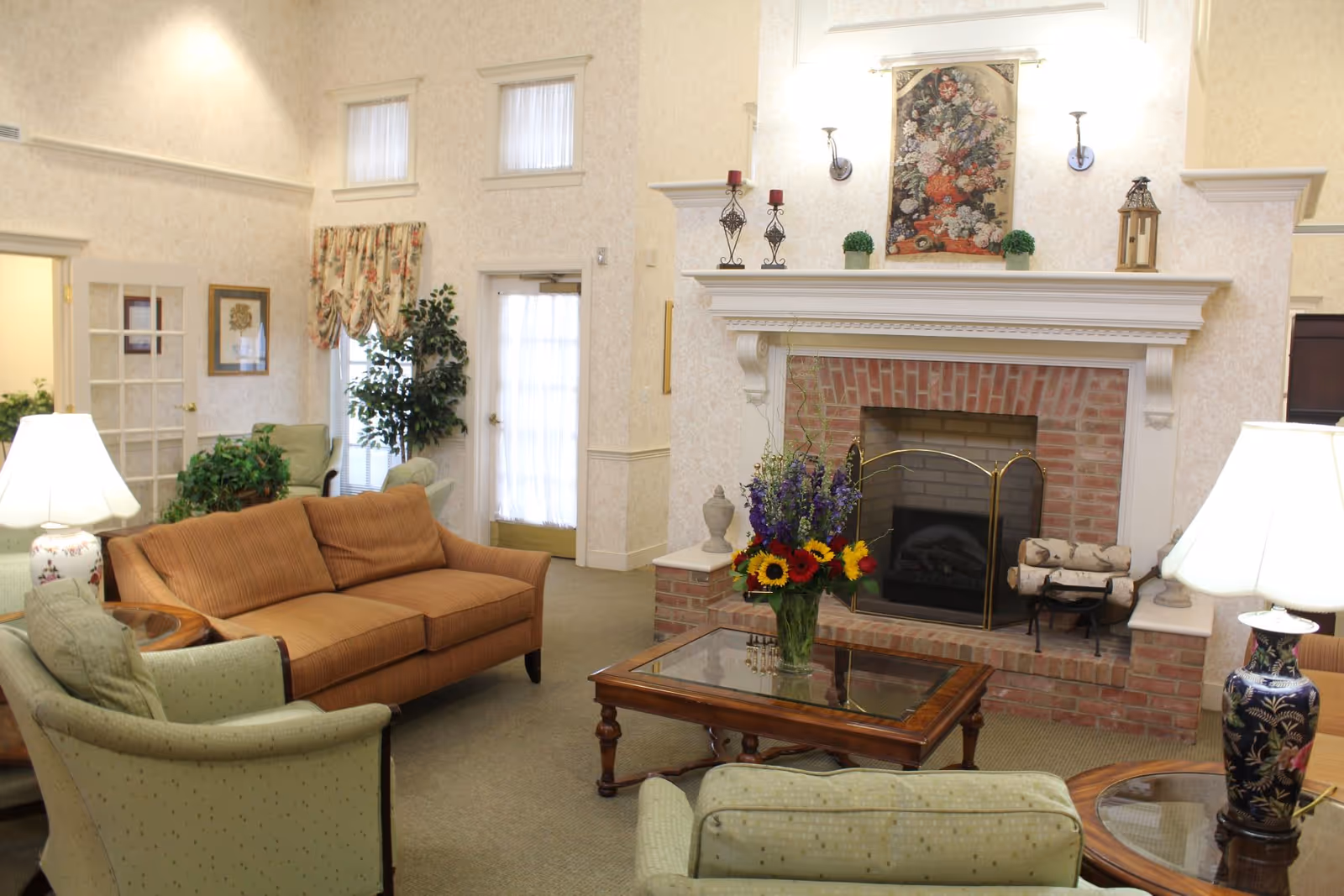 A cozy living room with a brick fireplace decorated with candles, plants, and a floral painting above it. The room has a brown sofa, two green armchairs, a glass-top wooden coffee table with a vase of colorful flowers, and two table lamps with floral-patterned bases. There are windows with floral curtains and a door with a sheer curtain in the background.