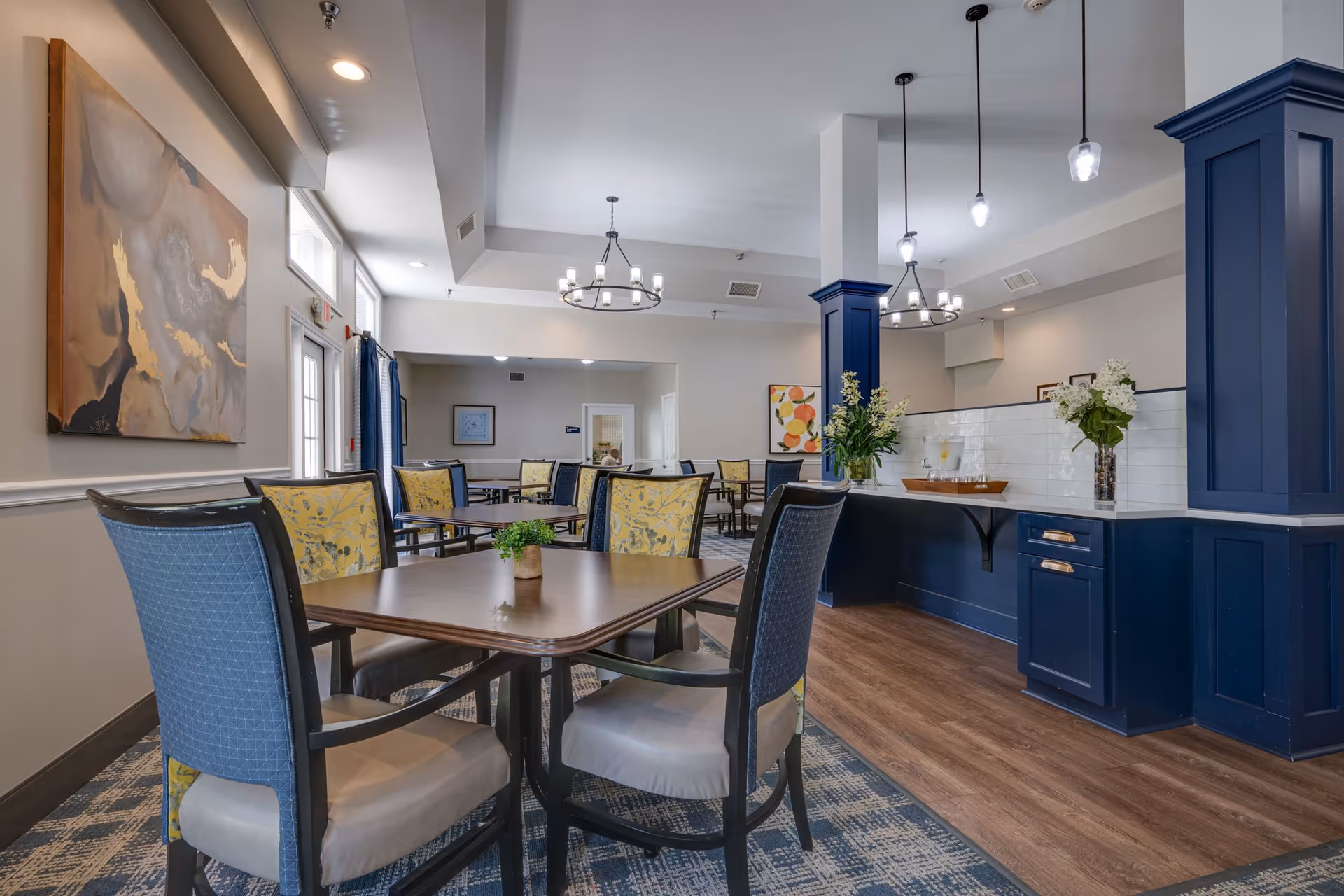 A bright and spacious dining room with multiple wooden tables and cushioned chairs featuring blue and yellow patterned upholstery. The room has modern chandeliers and pendant lights hanging from the ceiling. There is a blue cabinetry area with a white tiled backsplash and vases with flowers on the counter. The floor is a combination of wood and patterned carpet, and the walls are decorated with framed artwork.