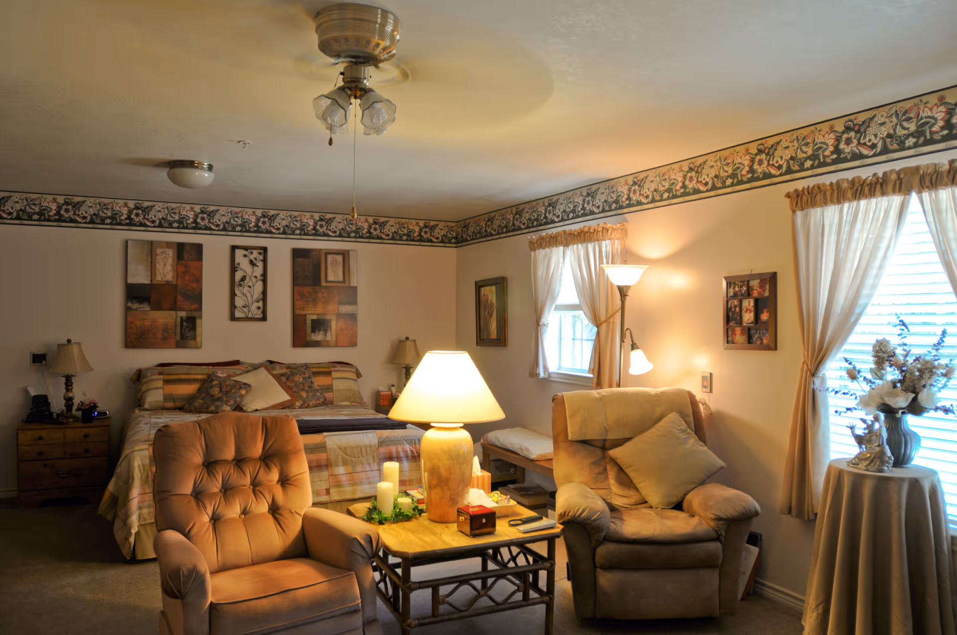 A cozy bedroom with a large bed against the wall, decorated with multiple pillows and a patterned bedspread. Two comfortable armchairs are positioned around a small wooden table with a lamp and candles. The room has floral wallpaper border near the ceiling, framed artwork on the walls, two windows with light curtains, and a floor lamp providing warm lighting.