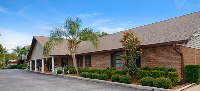 Single-story brick senior living building with a covered entrance, palm trees, and landscaped shrubs under a blue sky.