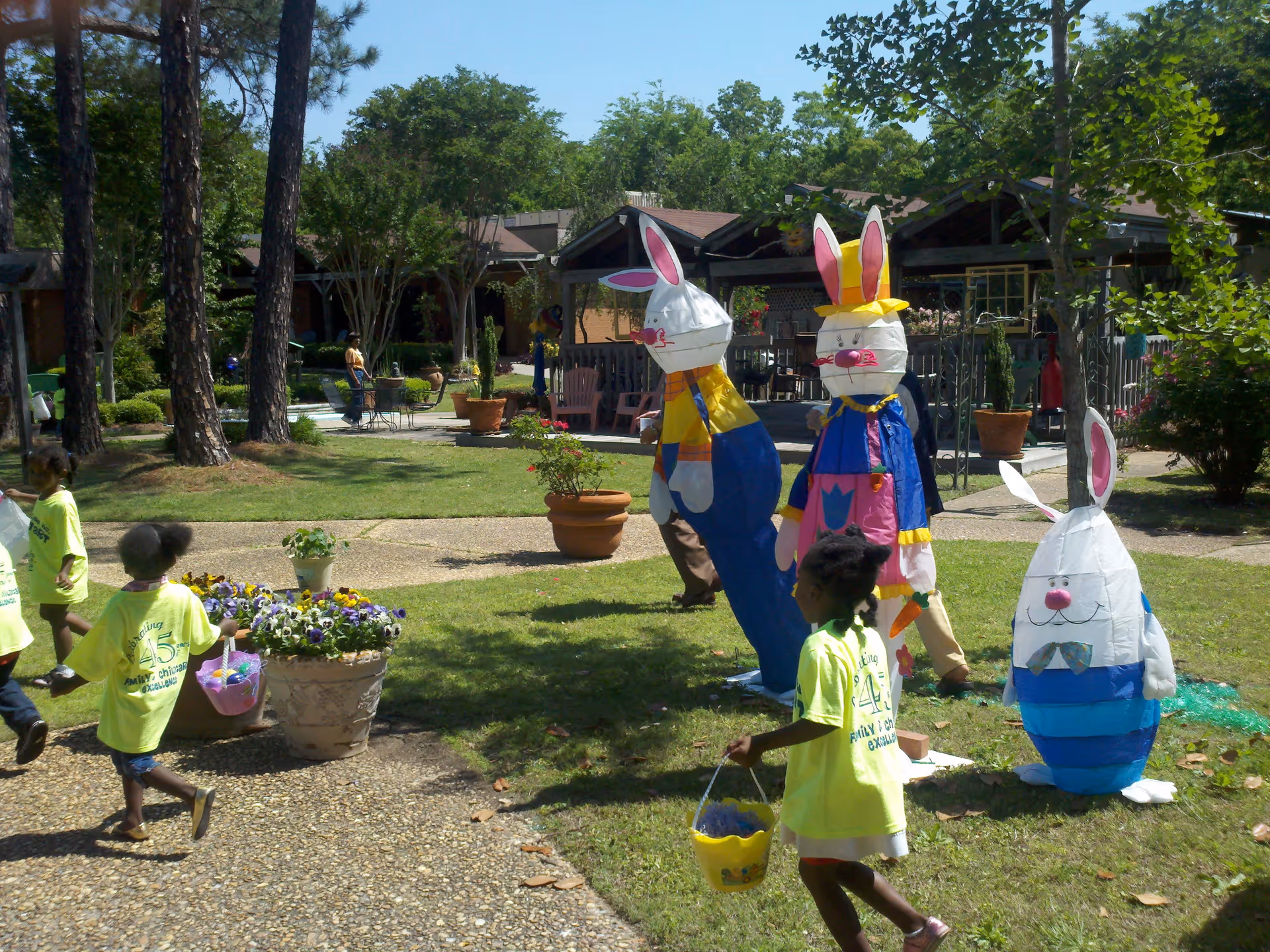 Children wearing bright yellow shirts are walking outdoors on a sunny day near large decorative Easter bunny figures in a garden area with trees, potted plants, and a building in the background.