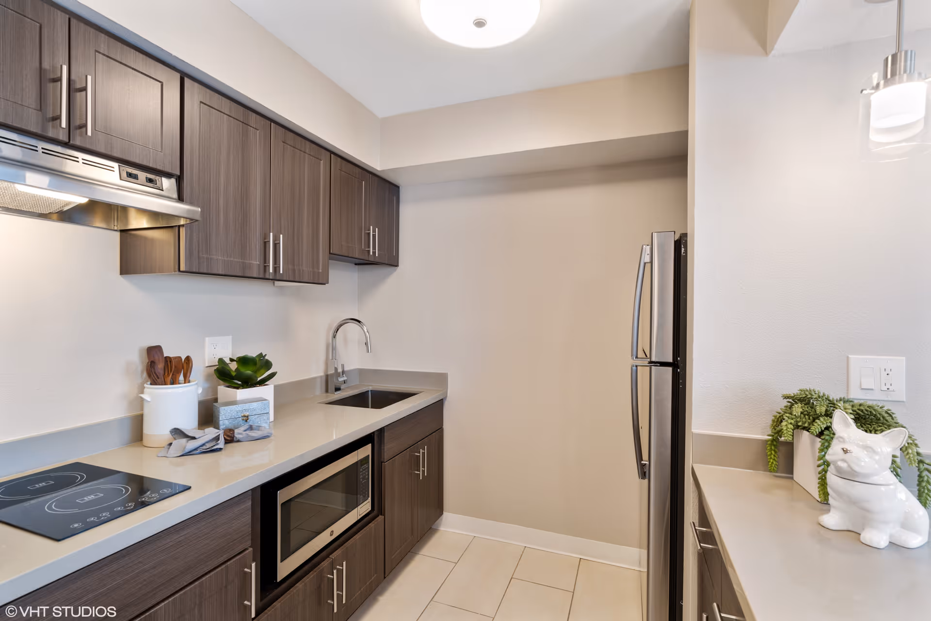 Modern kitchen with dark wood cabinets, a built-in microwave, an electric cooktop, a stainless steel refrigerator, a sink with a chrome faucet, and decorative items including a small plant and a white ceramic dog figurine on the countertop.