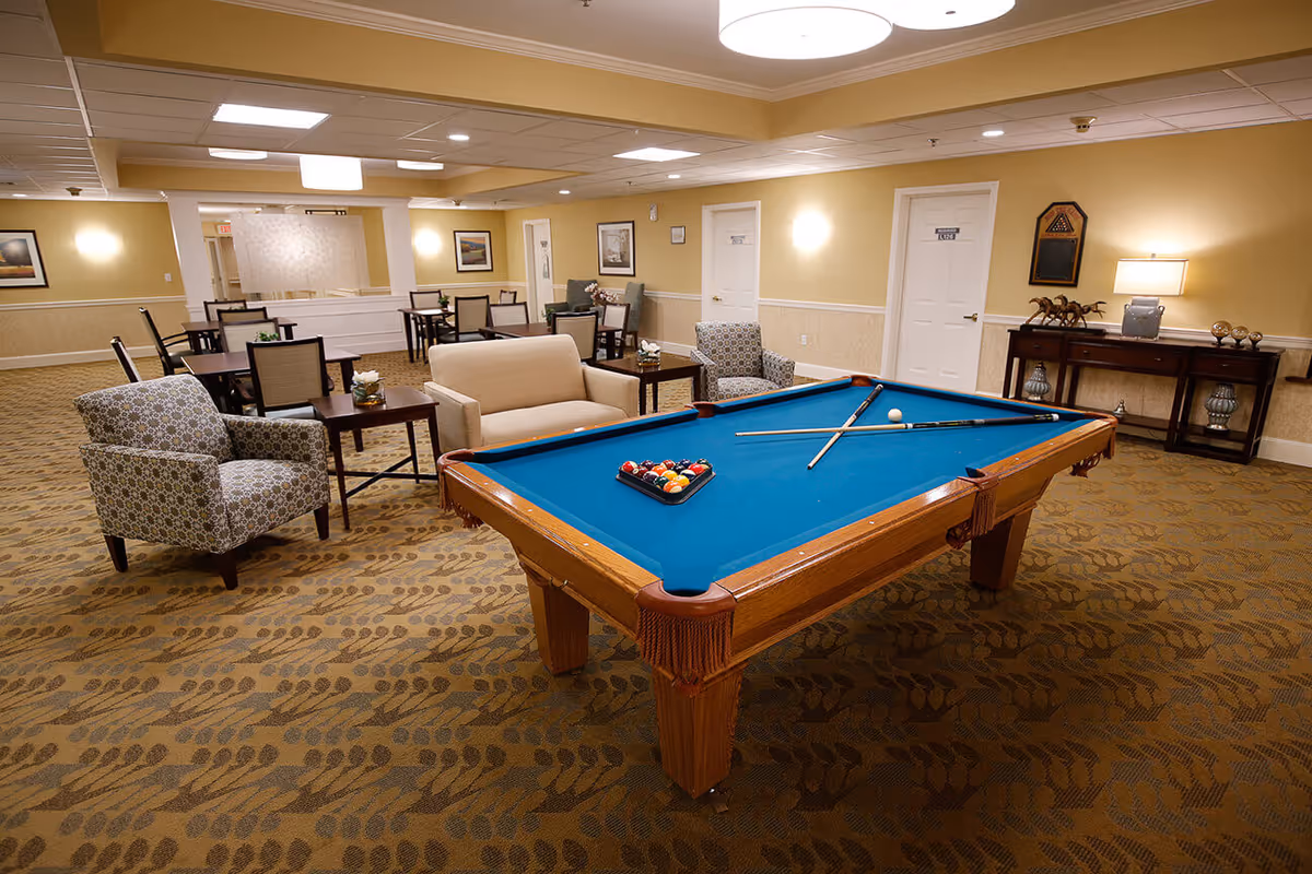A spacious common area in a senior living facility featuring a blue felt pool table with pool cues and balls arranged for a game. Surrounding the pool table are comfortable armchairs and a loveseat, with several tables and chairs arranged in the background. The room has beige walls, patterned carpet, and soft lighting from ceiling fixtures and wall sconces. A console table with decorative items and a lamp is against one wall.