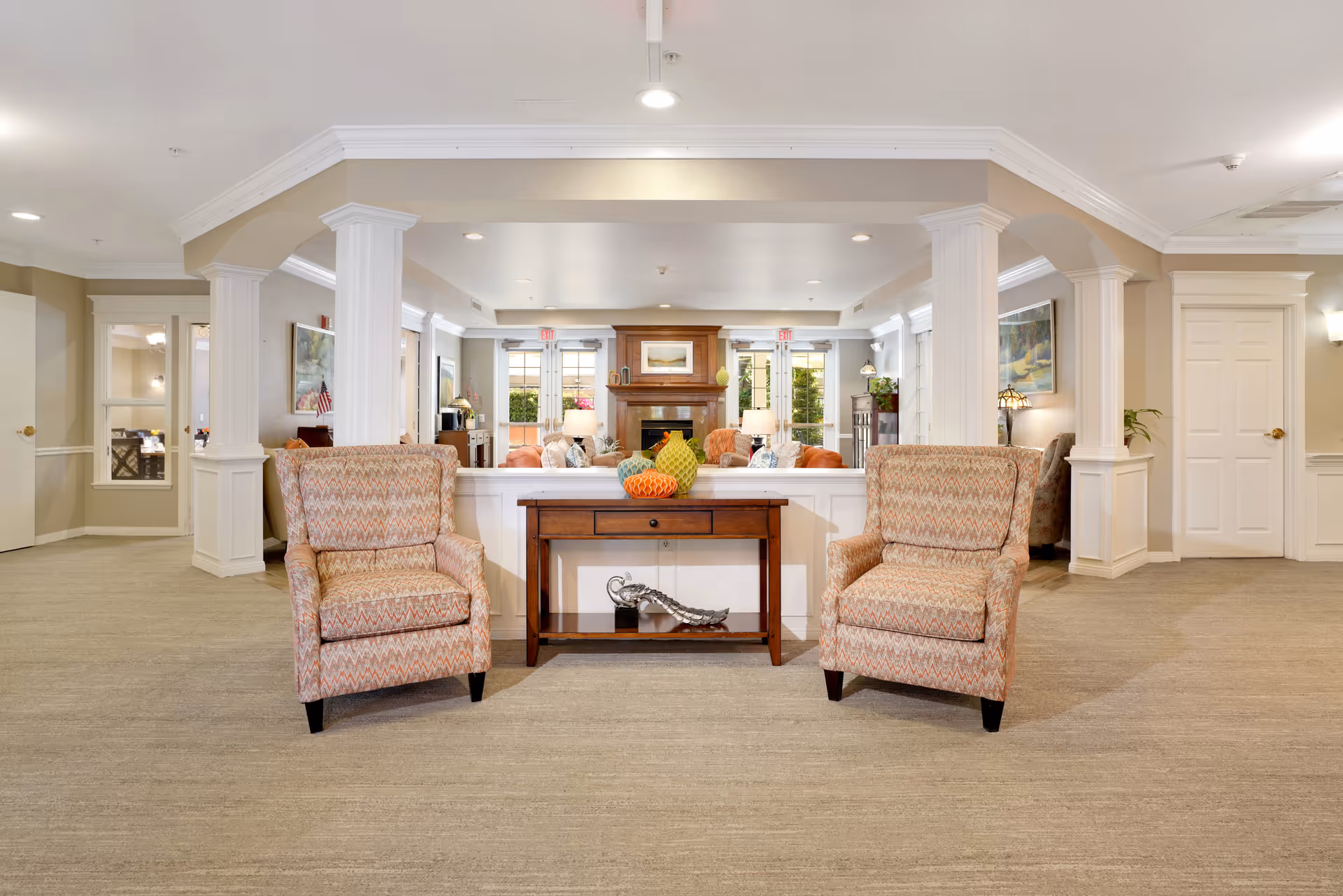 A spacious and well-lit living room area in a senior living facility featuring two patterned armchairs flanking a wooden console table with decorative items. The room has beige walls, white columns, and a fireplace in the background with seating arranged around it. Large windows and glass doors allow natural light to fill the space.