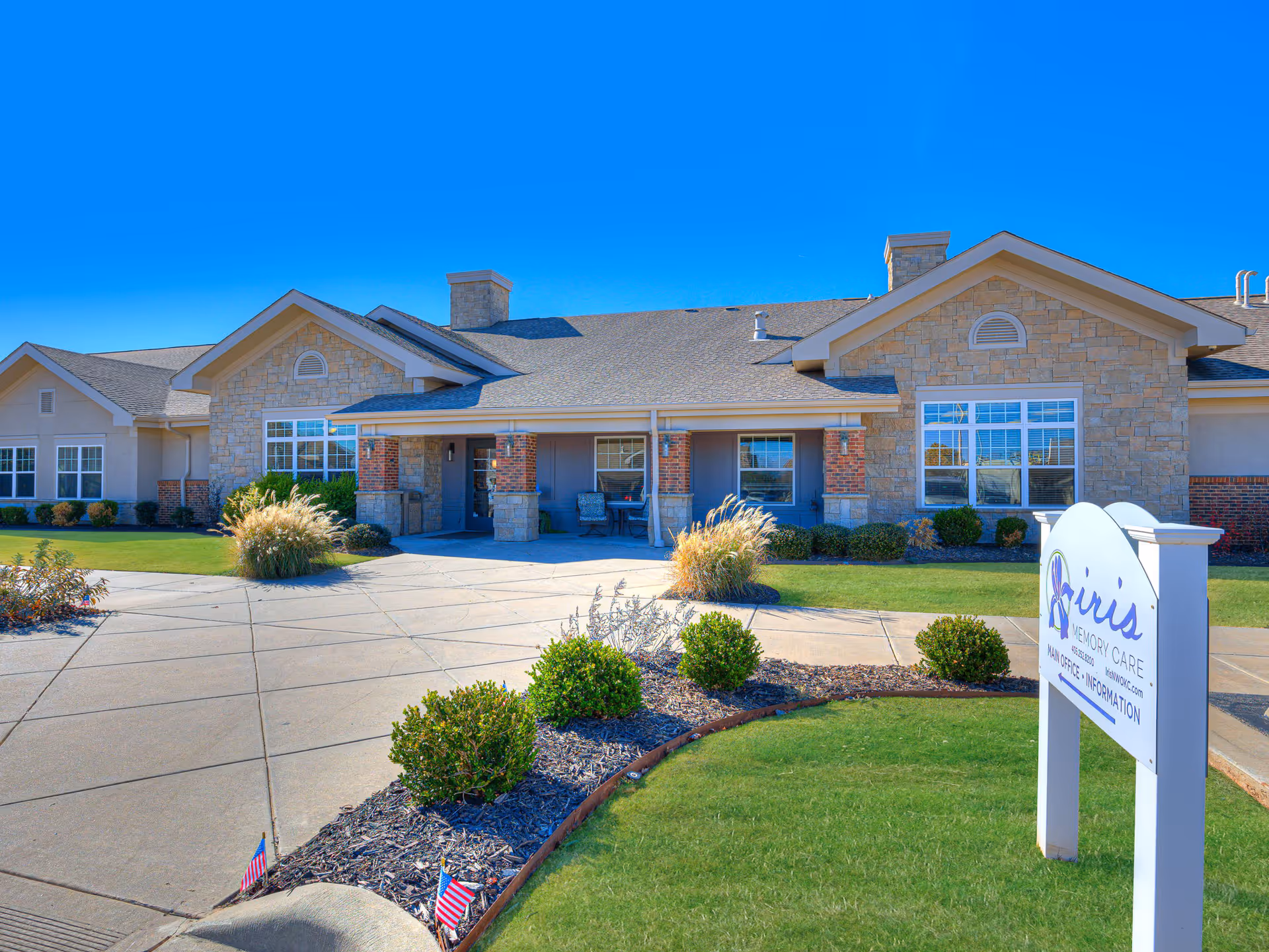 Exterior view of Iris Memory Care of NW Oklahoma City building with stone and brick facade, large windows, and a covered entrance. The foreground shows a well-maintained lawn, landscaped bushes, and a sidewalk leading to the entrance. A white sign with the facility name and contact information is visible on the right side.