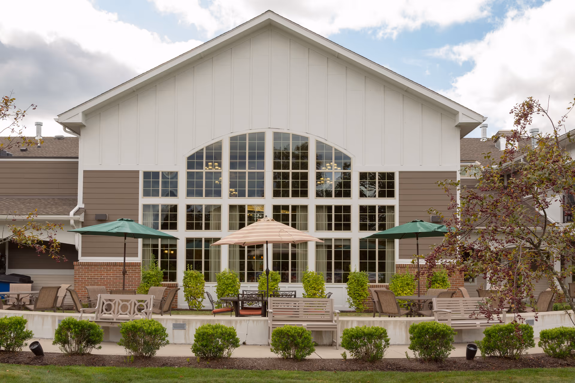 Front exterior of a senior living building with large arched windows and an outdoor patio area with umbrellas and seating.