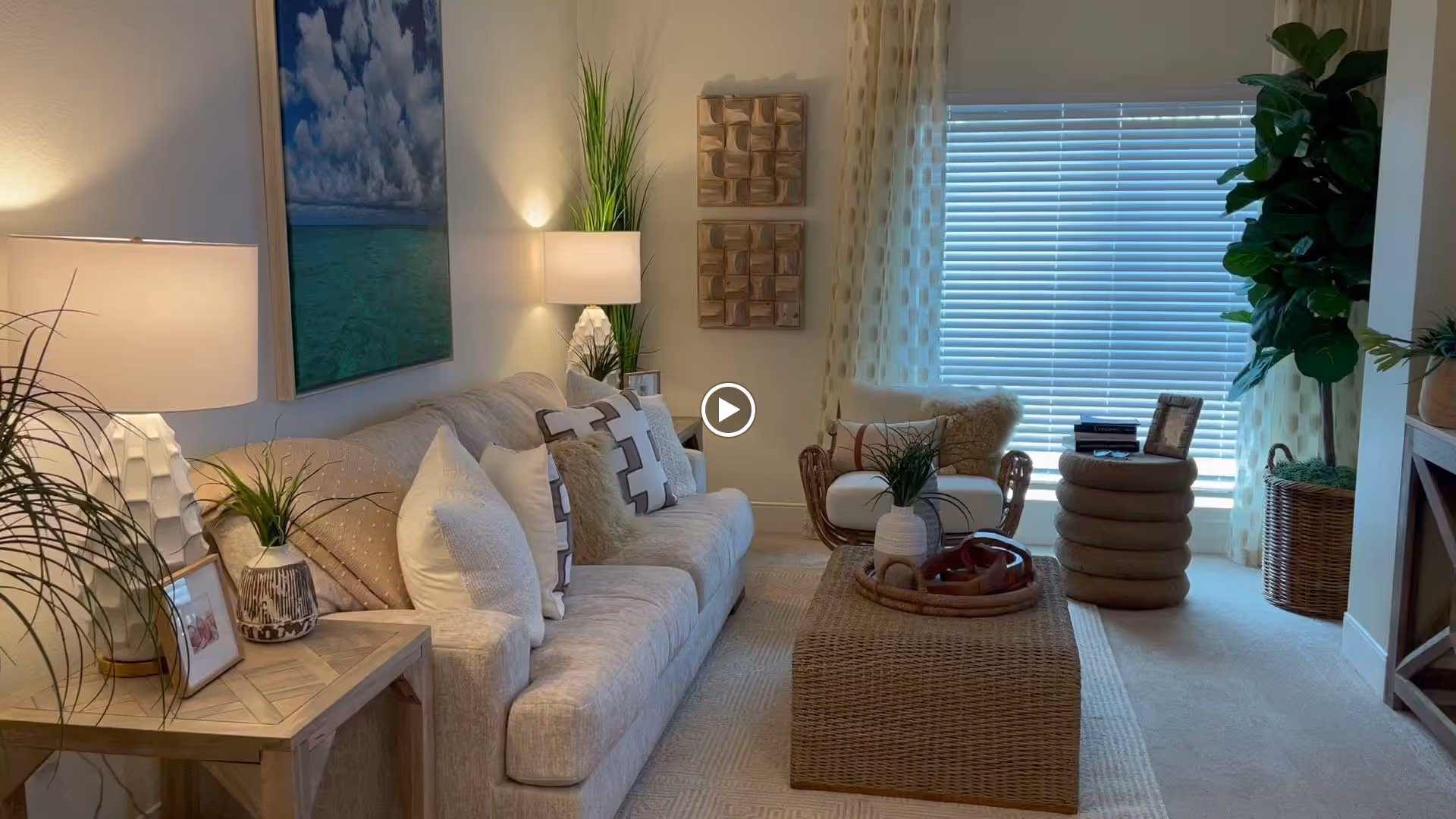 A cozy living room with a beige sofa adorned with multiple pillows, a wicker coffee table with decorative items, a cushioned chair, and a large window with blinds and patterned curtains. The room is decorated with plants, table lamps, wall art, and a light-colored carpet.