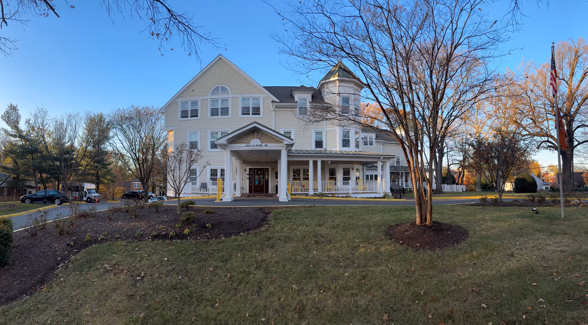 Front exterior of a white multi-story senior living building with a wraparound porch, trees, and a lawn.