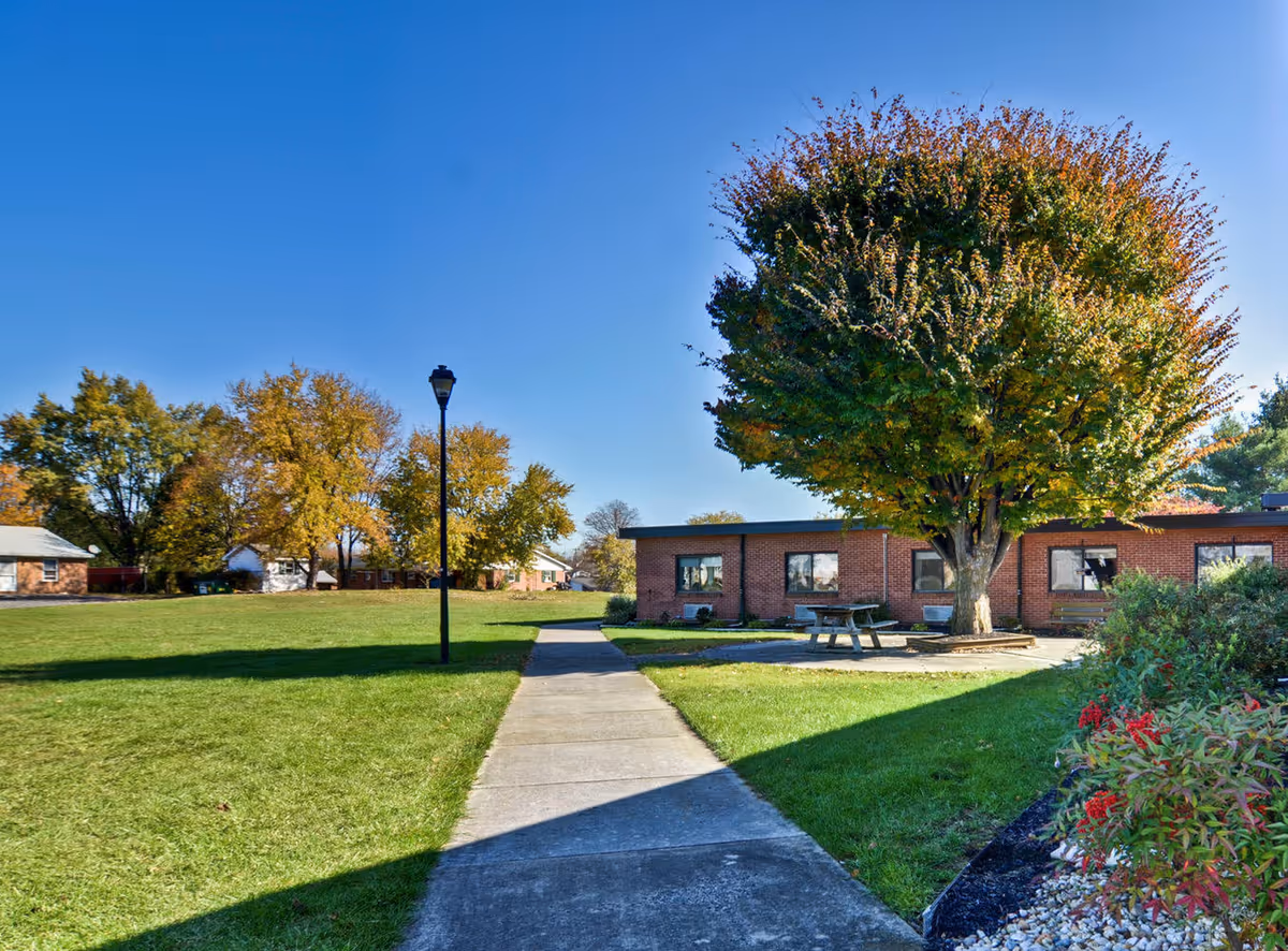 Outdoor view of Martinsburg Healthcare Center showing a paved walkway leading to a brick building with windows. There is a large tree with green and orange leaves near a picnic table, surrounded by grass and some bushes with red berries. The sky is clear and blue.