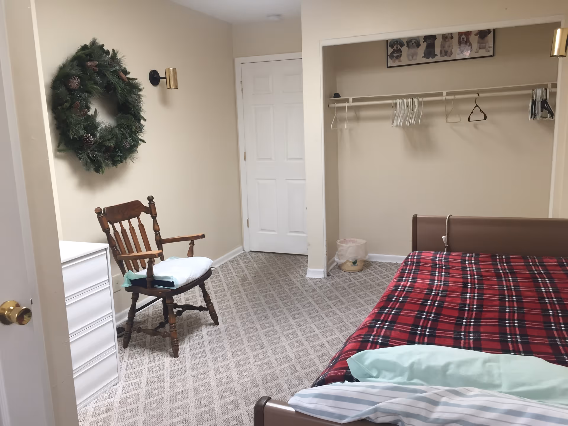 Small furnished bedroom with a single bed covered in a red plaid blanket, a wooden chair, dresser, open closet and a wreath on the wall.