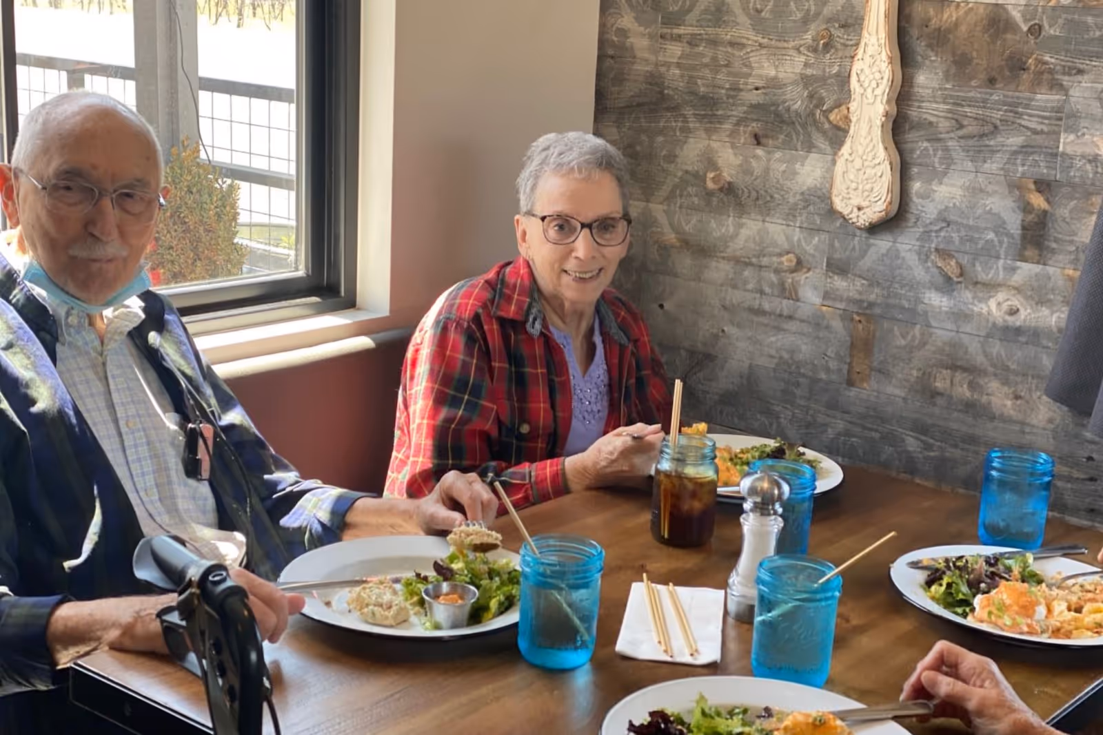 Two elderly people sitting at a wooden dining table near a window, enjoying a meal with plates of food and blue drinking glasses. The wall behind them is made of rustic wood panels with a decorative hanging piece.