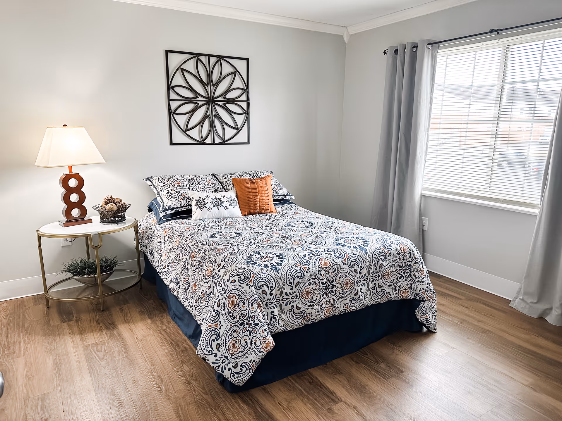 A neatly made bed with patterned bedding and decorative pillows in a bright bedroom with light gray walls, a window with gray curtains, a round side table with a lamp and decorative items, and a metal wall art piece above the bed.