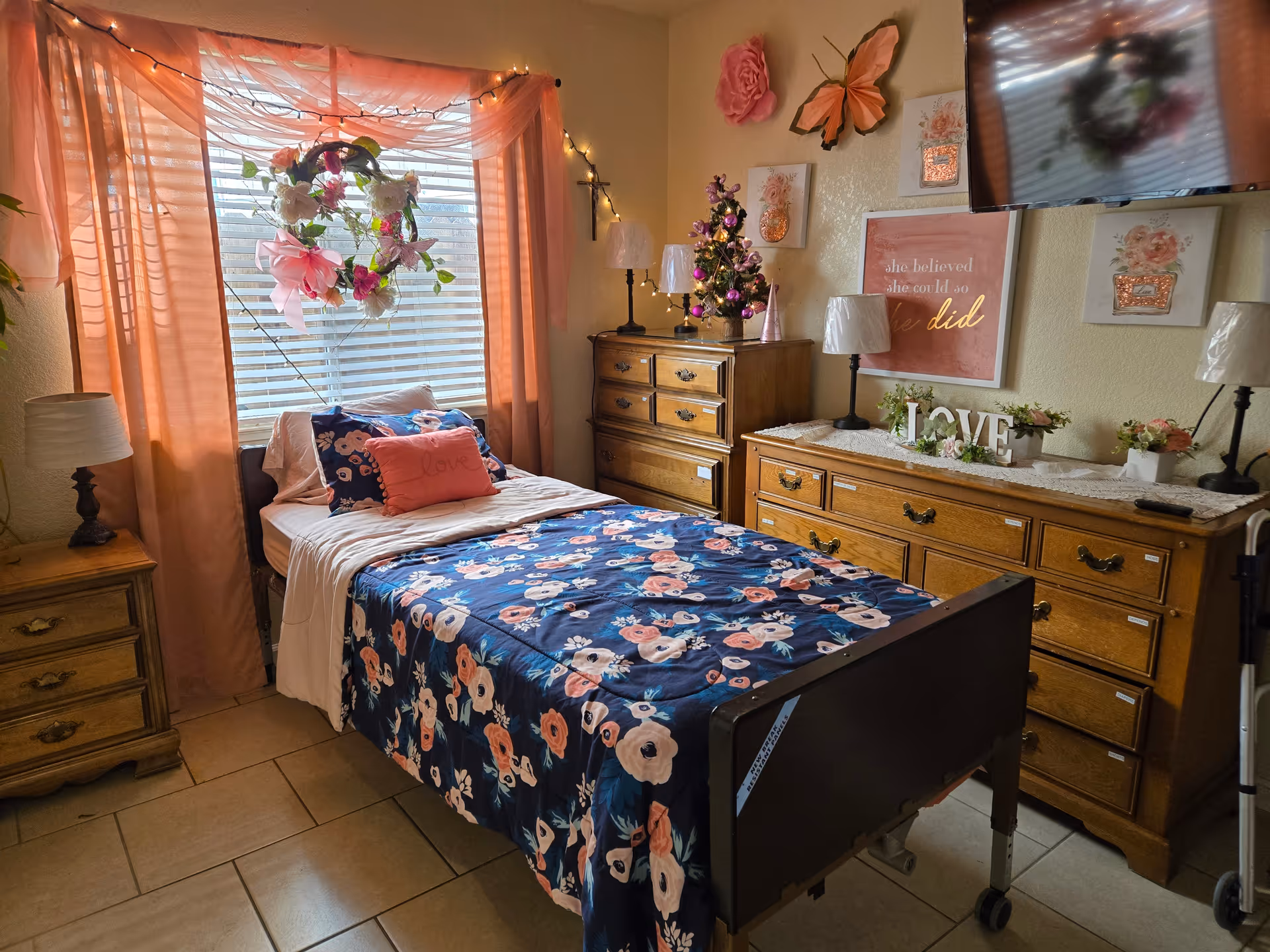 A cozy bedroom in Siebenthal Elder Care Home featuring a single bed with floral bedding and pillows, a window with pink curtains and a floral wreath, wooden dressers with lamps and decorative items including a small Christmas tree, wall art, and a mounted TV.