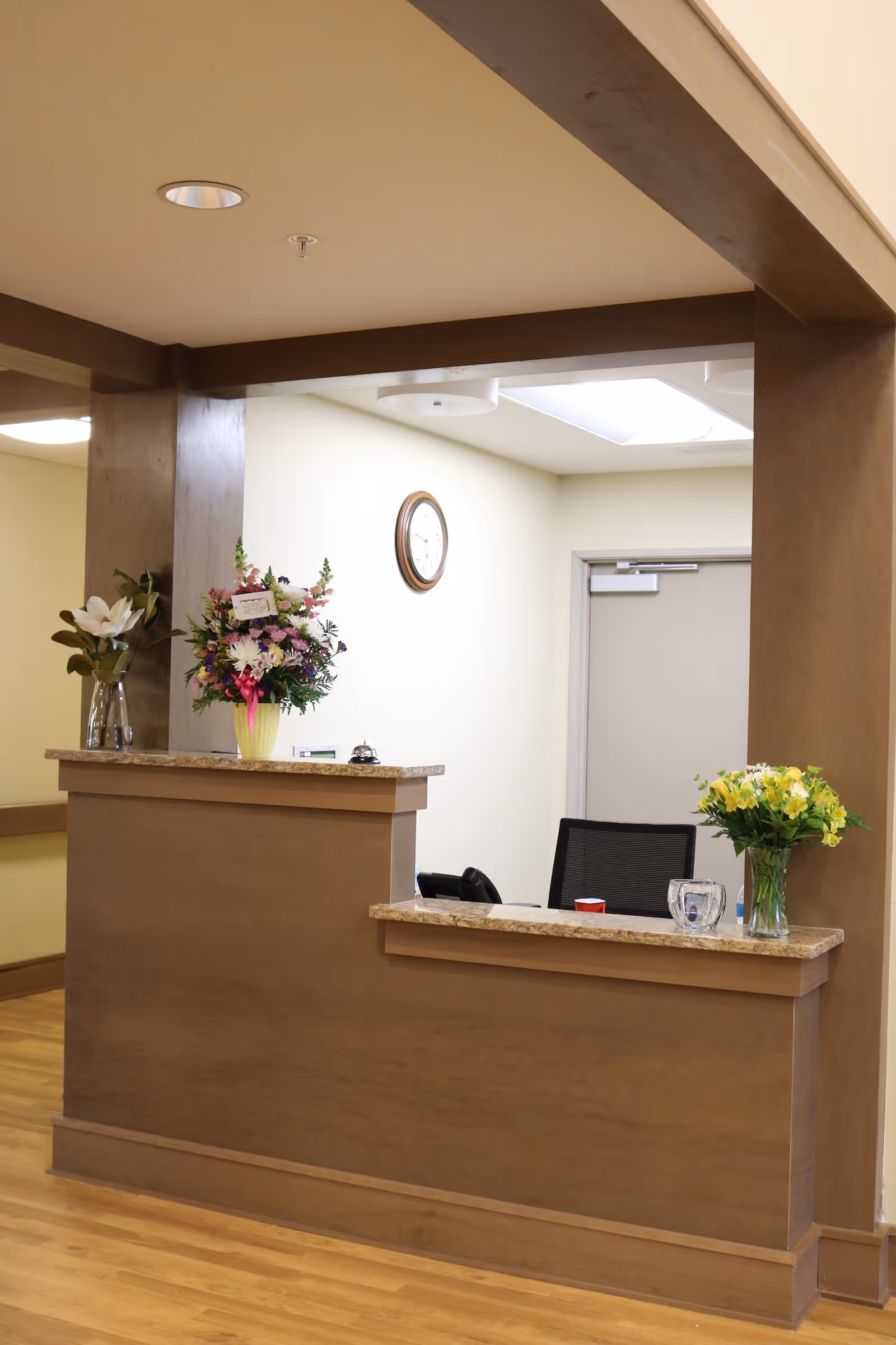 Reception desk area in a senior living facility with a wooden counter, three vases of flowers, a clock on the wall, and a closed door in the background.