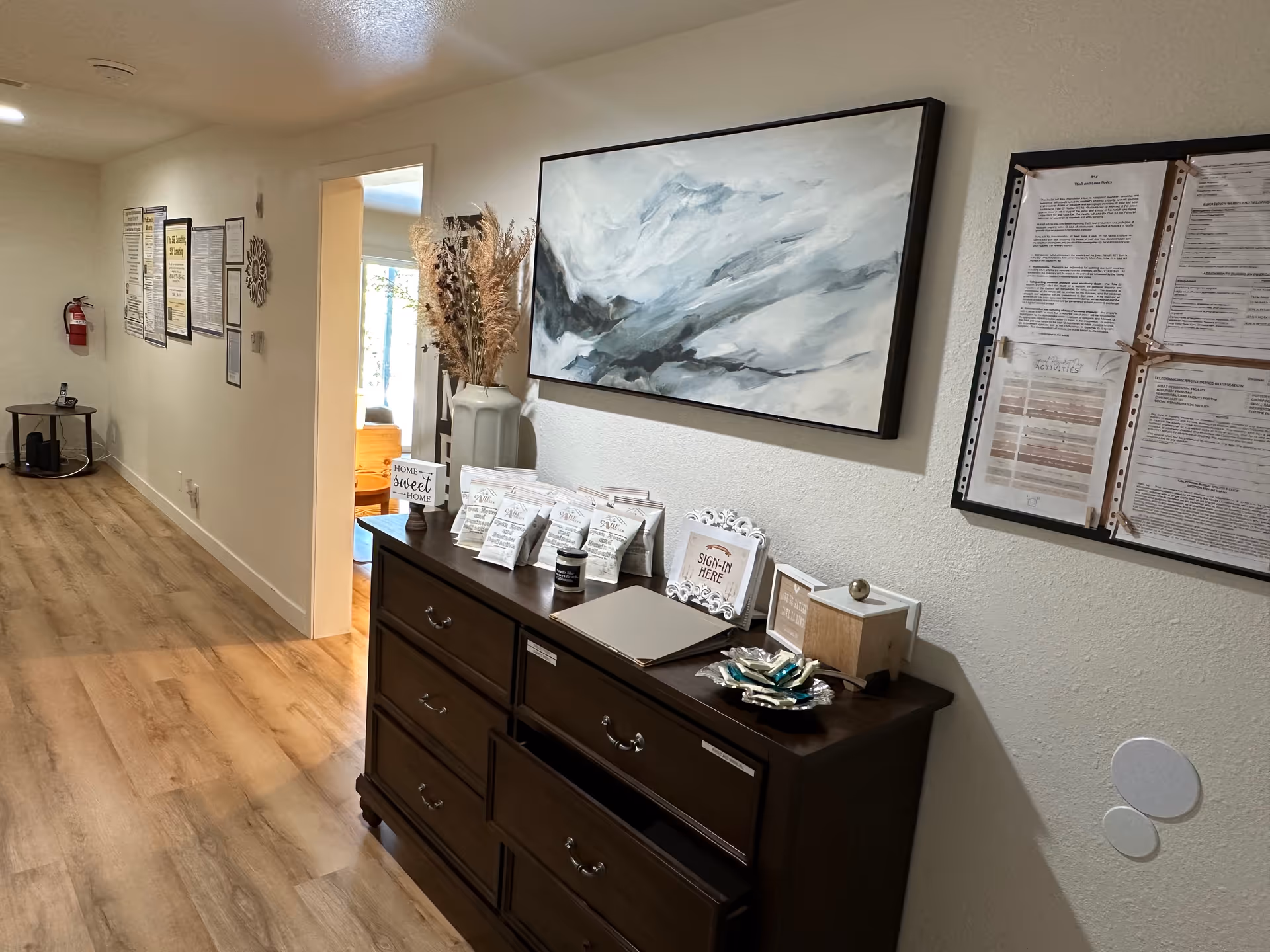 Interior hallway of a senior living facility with light wood flooring and white walls. A dark wooden dresser is against the right wall, decorated with a large abstract painting above it, a vase with dried plants, a sign that says 'Home Sweet Home,' a sign-in book, and various small items. On the wall above the dresser are several framed documents and notices. The hallway extends to the left with more framed papers on the wall and a fire extinguisher mounted near the end.