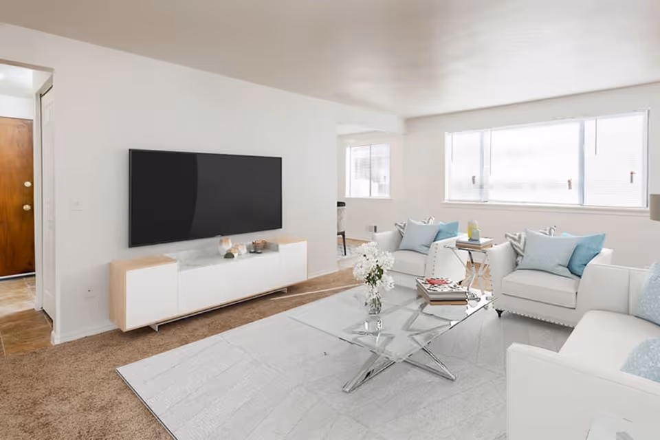 Bright and modern living room with white walls, a large flat-screen TV mounted on the wall above a wooden and white media console, a glass coffee table with a vase of white flowers, and white sofas and armchairs with light blue and patterned throw pillows. Large windows allow natural light to fill the room.