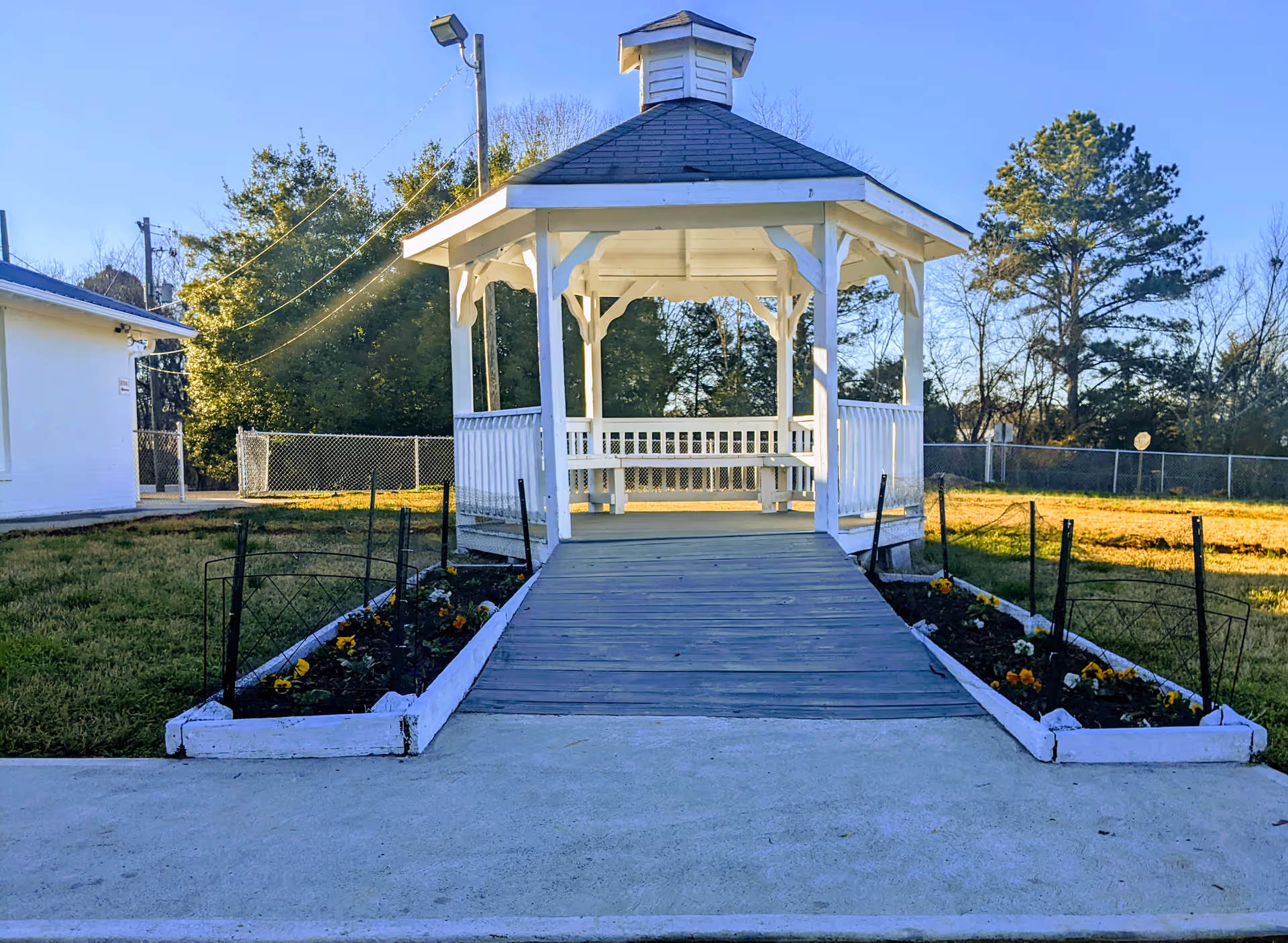 White wooden gazebo with a ramp and flower beds in a fenced grassy yard under a clear blue sky.
