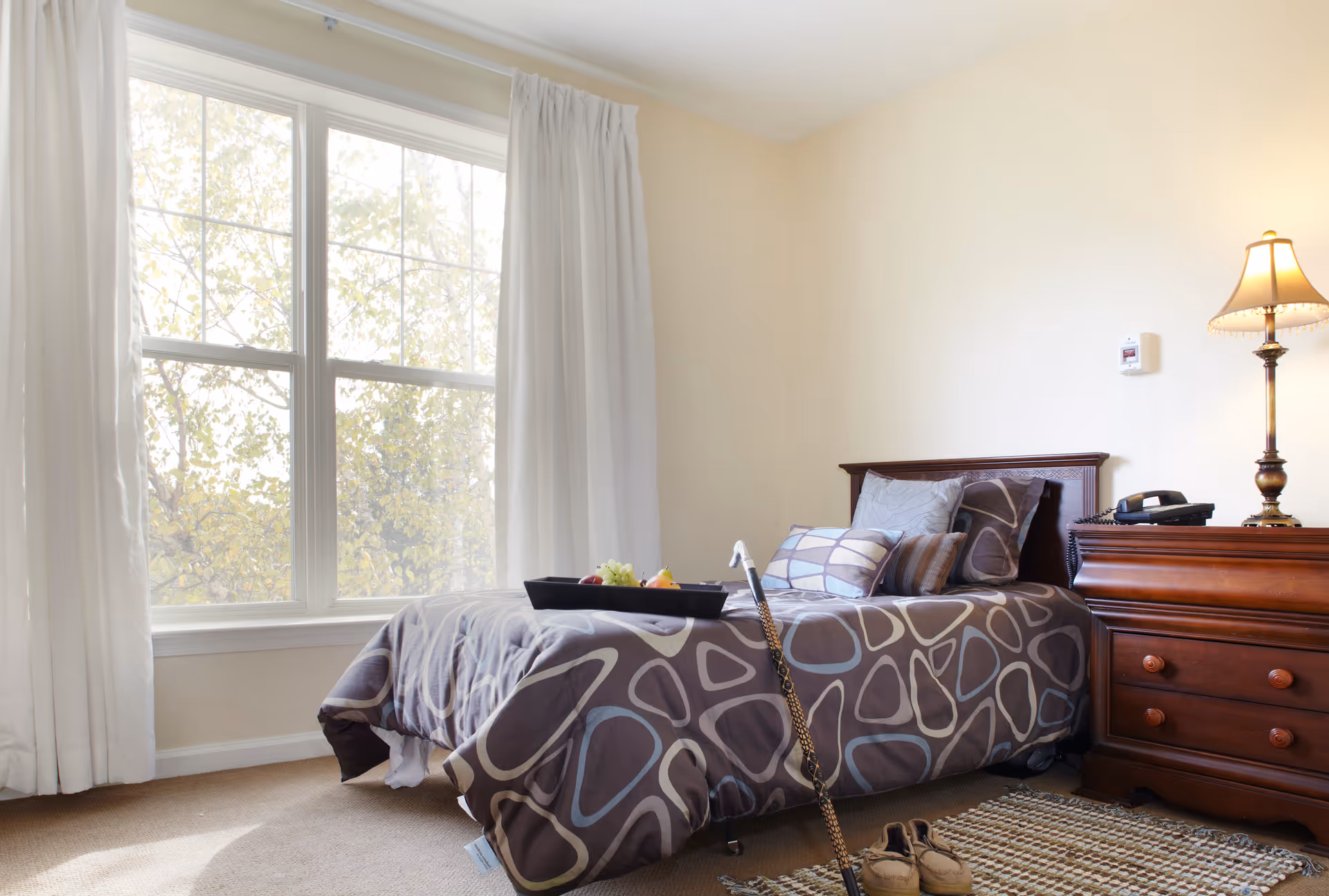 Sunlit bedroom with a single bed dressed in patterned bedding, a wooden nightstand with a lamp, a cane and slippers on the floor.