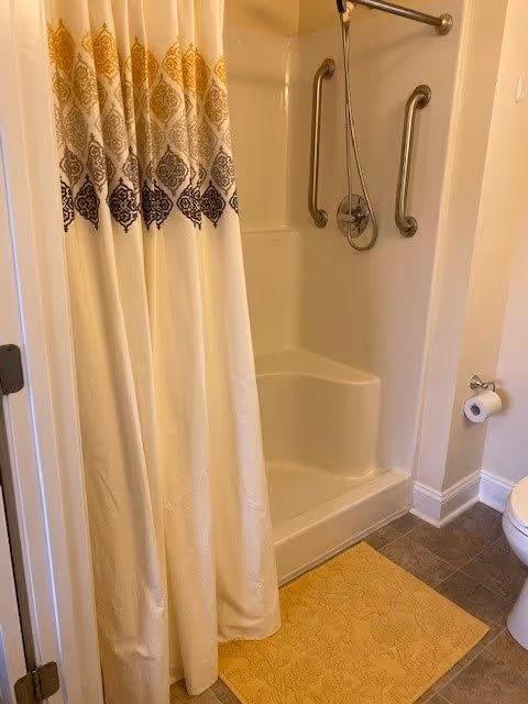 A bathroom shower area with a white and patterned shower curtain partially drawn. The shower has built-in seating and two metal grab bars for support. A handheld showerhead is mounted on the wall. A yellow bath mat is placed on the tiled floor outside the shower. A toilet paper holder with a roll of toilet paper is visible on the right side.