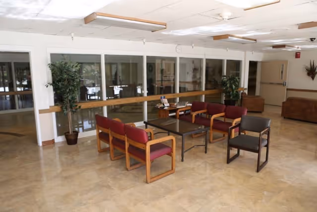 A seating area in a senior living facility with several wooden chairs with red cushions arranged around a rectangular coffee table. There are potted plants near the glass windows and doors in the background, and the floor is covered with beige tiles. The ceiling has fluorescent lights and there is a hallway visible to the right.