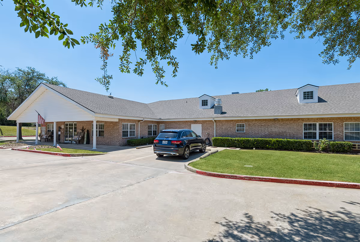 Exterior view of a single-story brick building with a gray shingled roof, a covered entrance with white pillars, an American flag, and a black SUV parked in front. The building is surrounded by green grass and trimmed bushes under a clear blue sky with tree branches hanging overhead.