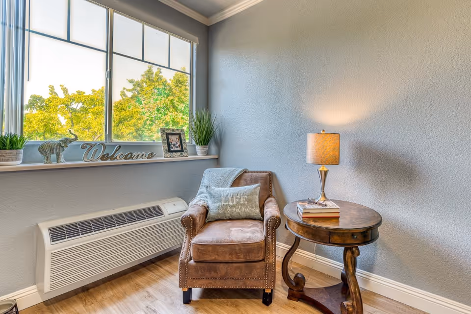 Cozy corner seating area with a brown leather armchair, round wooden side table with a lamp, and a window sill decorated with plants and a 'Welcome' sign.
