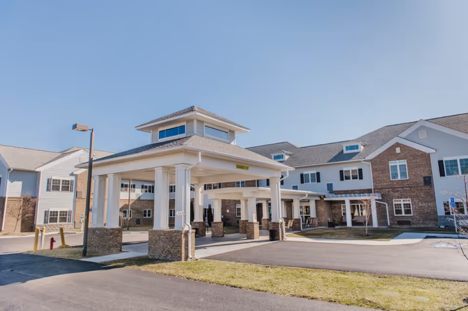 Exterior view of English Meadows Stephens City Campus showing a large covered entrance with white columns and brick bases, part of a multi-story building with white siding and brick accents under a clear blue sky.