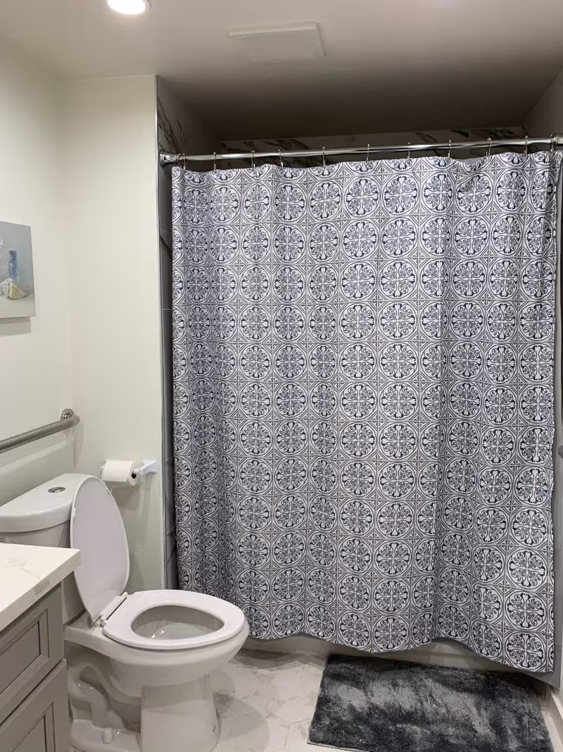 A bathroom with a white toilet with the lid open, a toilet paper holder mounted on the wall, a patterned shower curtain with a circular design, a gray bath mat on the floor, and a portion of a vanity with a marble countertop visible on the left side.
