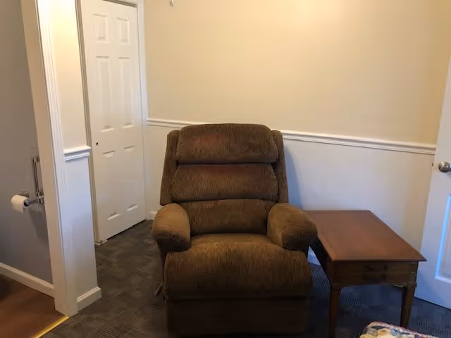 A brown upholstered recliner chair positioned next to a wooden side table with a drawer in a room with beige walls and white trim. There is a white door with a silver knob on the right and a closet with double white doors in the background. A toilet paper holder is mounted on the left wall near the doorway.