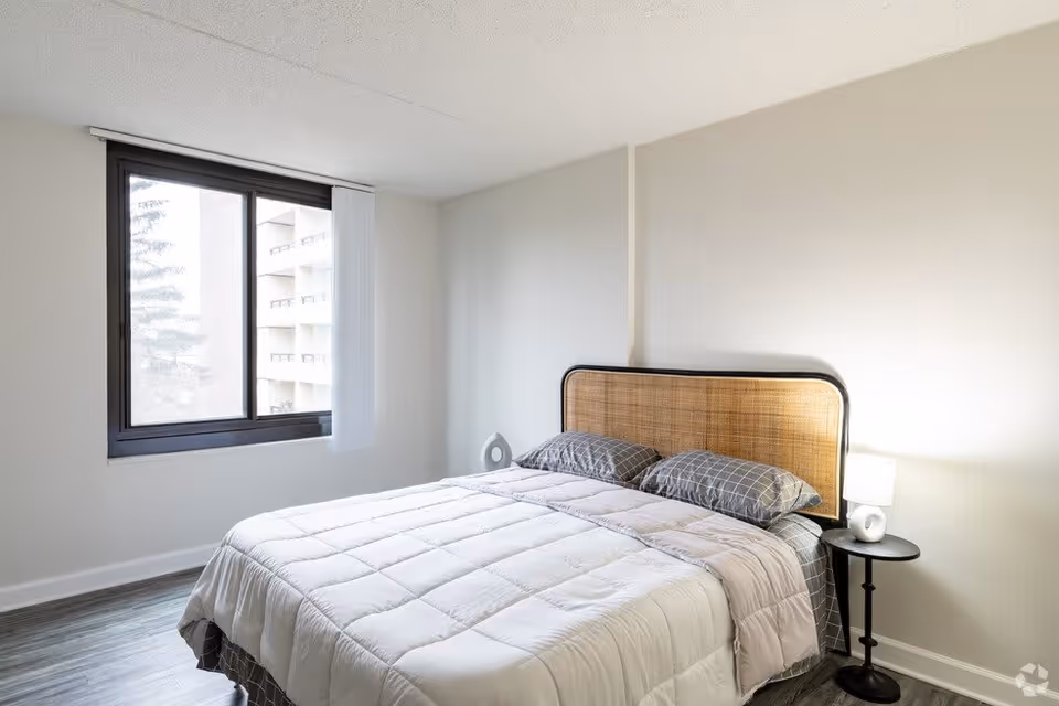 A bright bedroom with a large window, a bed with a woven headboard, gray checkered pillows, a light gray comforter, and a small round black bedside table with a modern lamp and decorative object.